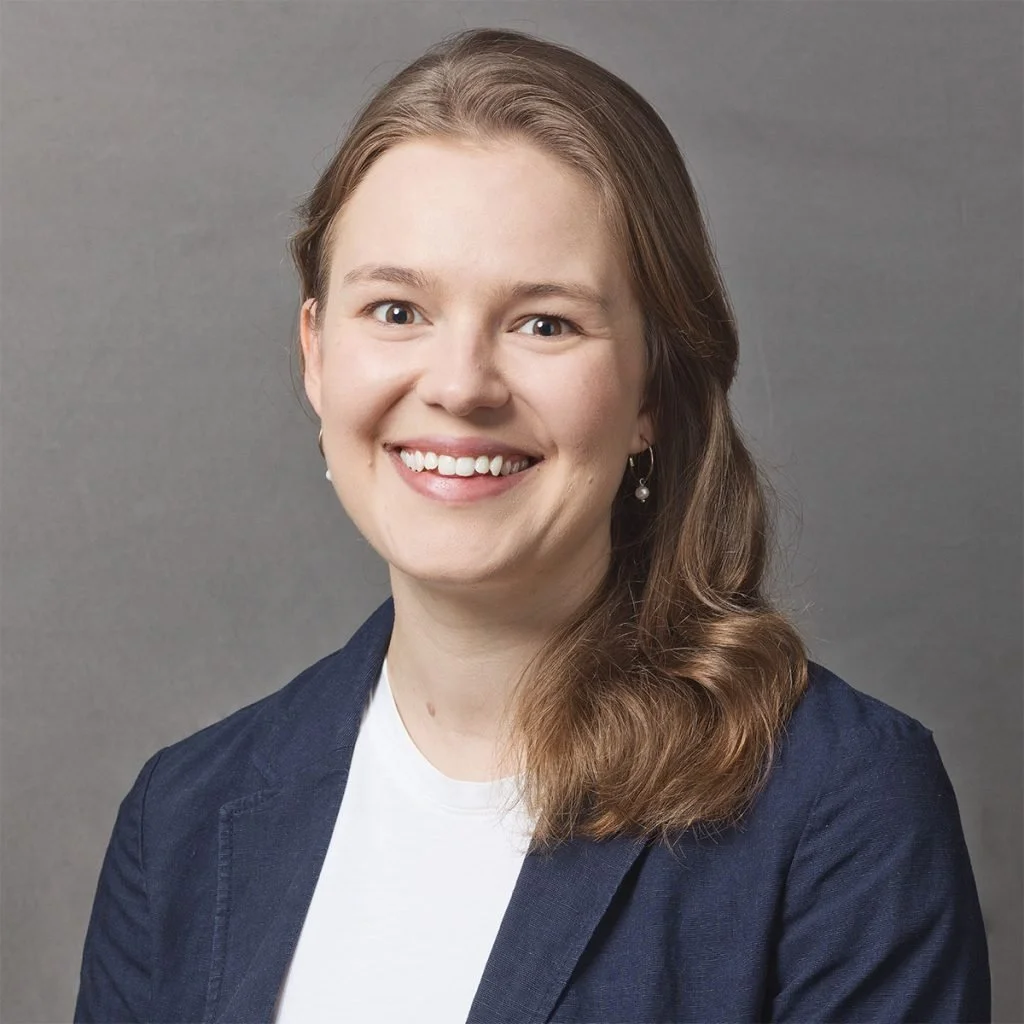 A woman with light skin, long brown hair, and earrings, smiling, wearing a navy blazer over a white top, against a plain gray background.