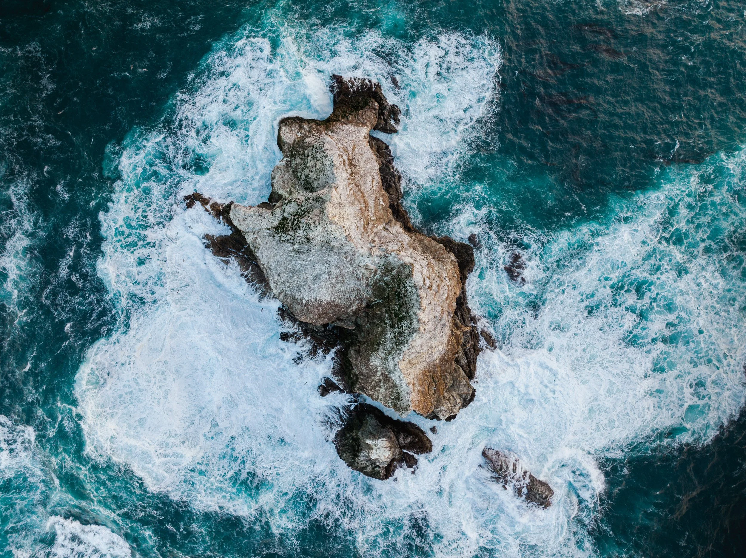 Bird's-eye view of a large rock formation in the ocean surrounded by turquoise water and crashing white waves.