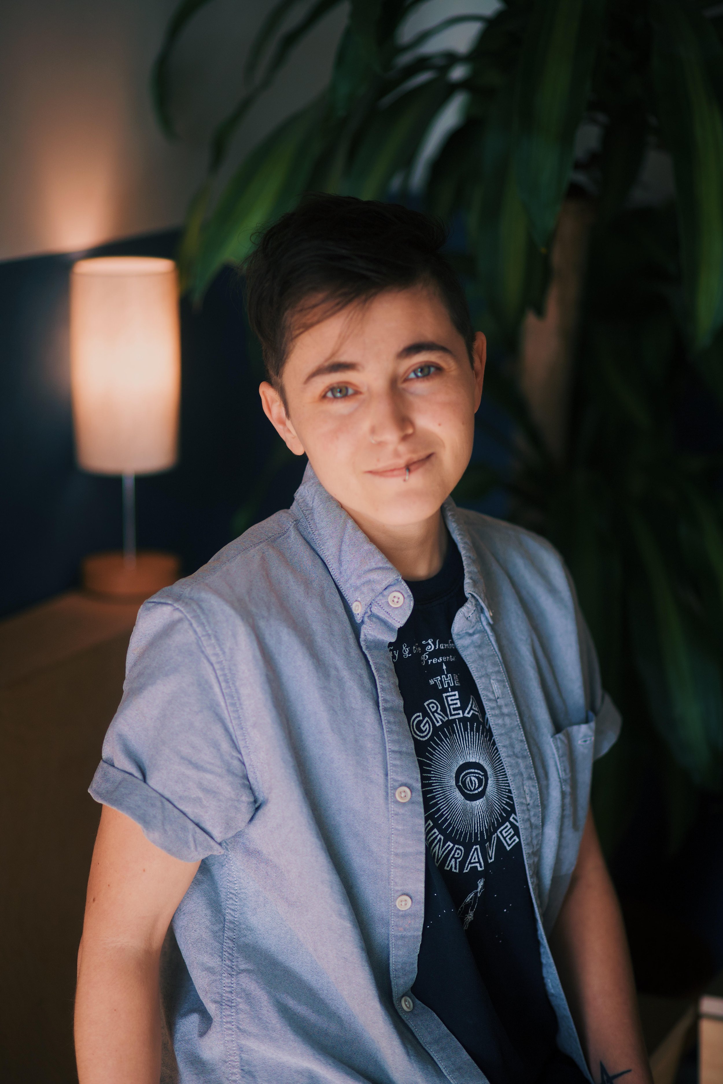 A young person with short dark hair and piercing blue eyes, smiling slightly, seated indoors next to a lamp and large leafy plant, wearing a casual shirt over a graphic T-shirt.