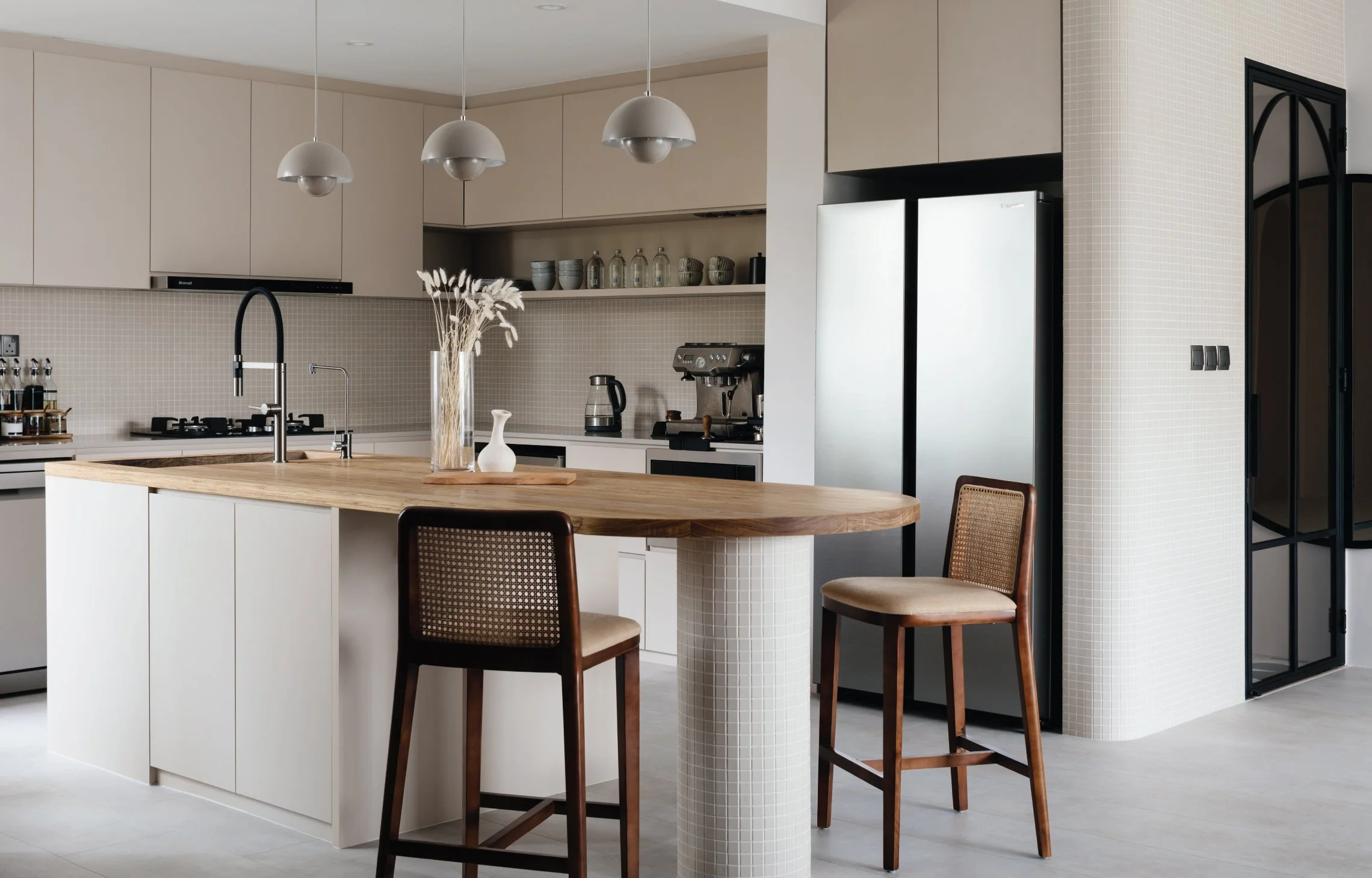 Modern kitchen with beige cabinets, a wooden island with a sink, and three wooden chairs with woven backs. There are pendant lights hanging from the ceiling, a vase with dried flowers on the island, and kitchen appliances like a coffee machine and refrigerator.