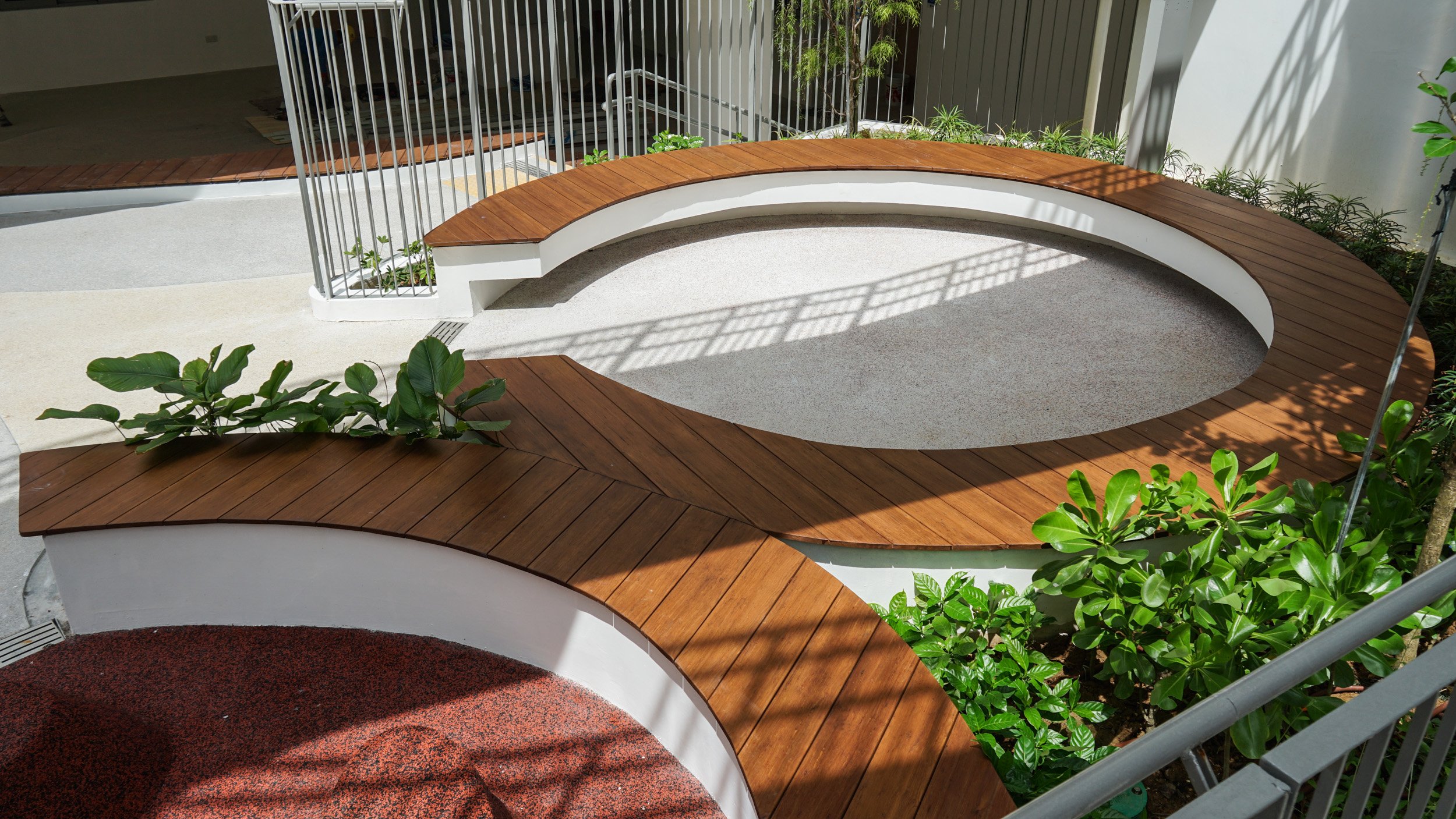Outdoor ONEWOOD seating area with curved wooden benches and green plants, surrounded by white walls and a metal railing.