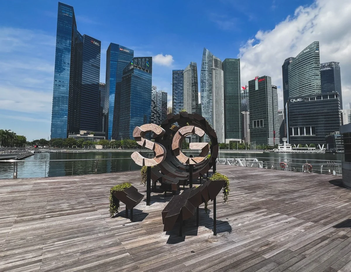 Modern city skyline with tall glass skyscrapers and a waterfront promenade. A large sculpture with the letters 'SG' is in the foreground on the wooden deck.