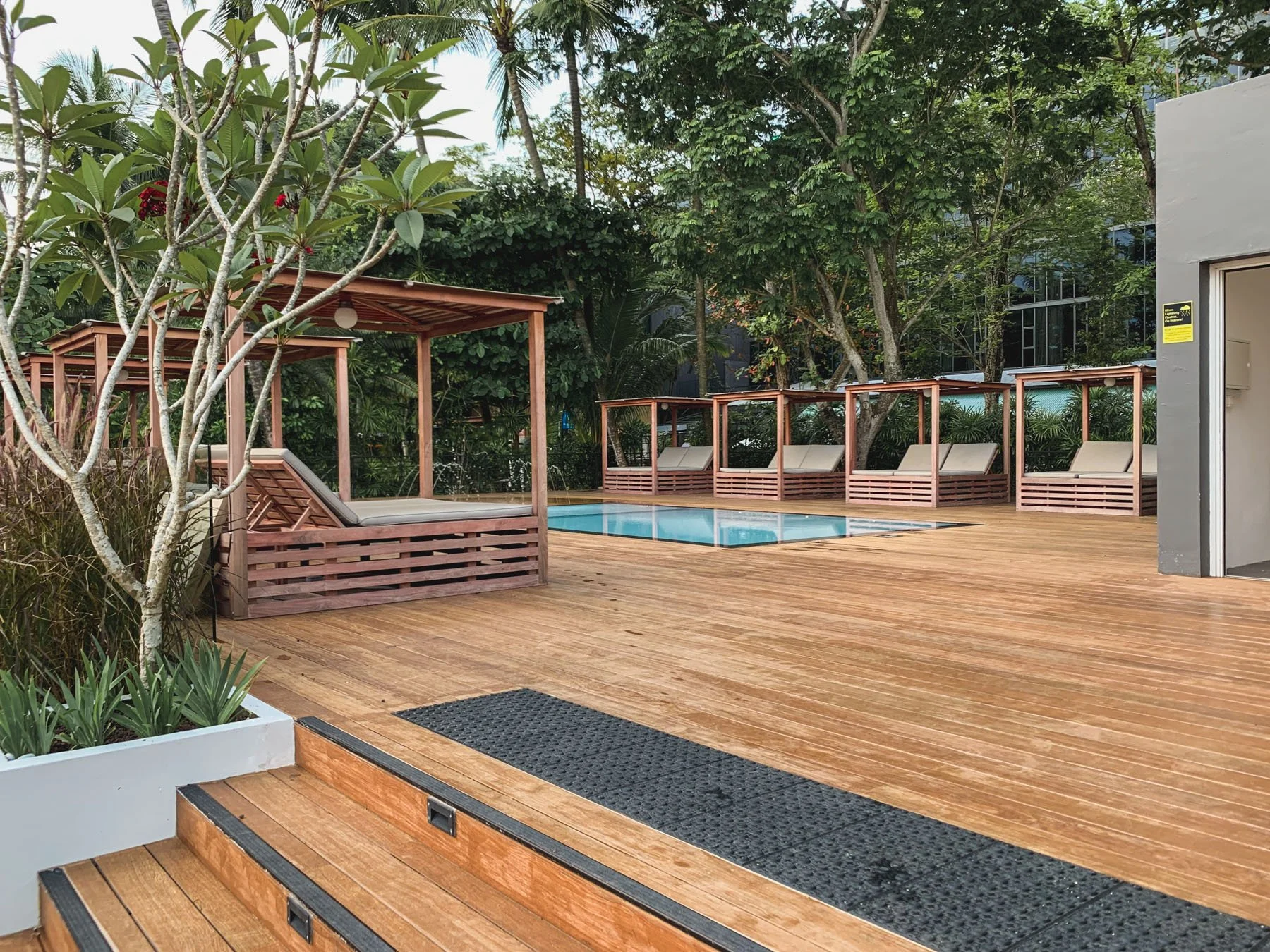 An outdoor pool area with ONEWOOD Timber decking, several private cabana-style lounge beds with beige cushions, surrounded by lush green trees and plants.