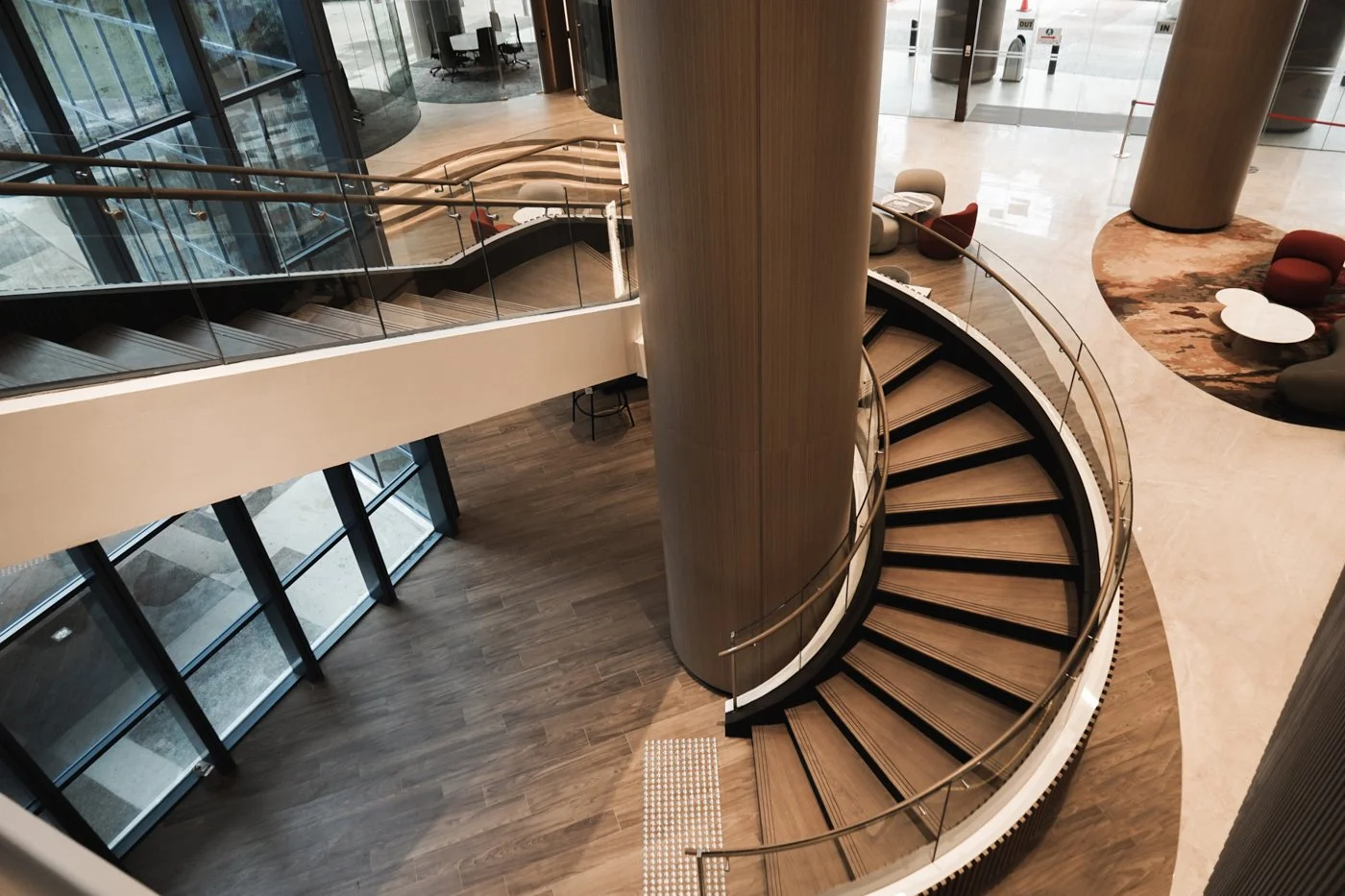Interior view of a modern building with a spiral staircase, wooden floors, and seating areas with chairs and tables near large glass windows.