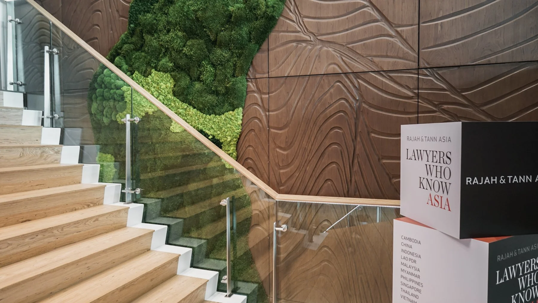 Onewood Timber Interior staircase with wooden steps and glass railing, next to a green moss wall and a wooden textured wall with a carved wave pattern.
