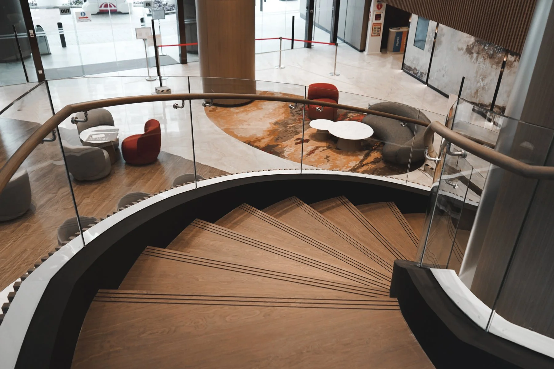 Onewood Solid Timber spiral staircase with wooden steps and glass railings, overlooking a seating area with chairs, tables, and a patterned rug, near a large glass entrance in a hotel lobby.