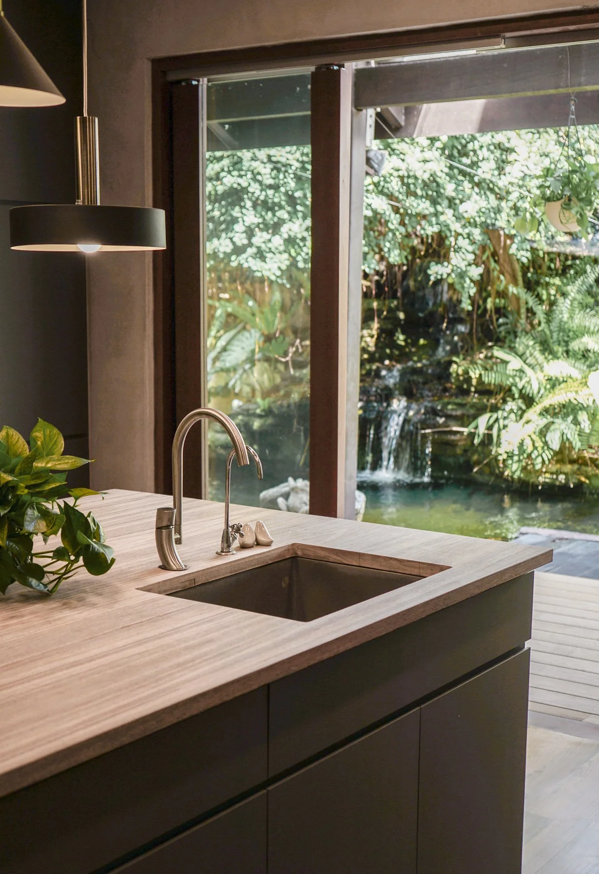 Modern kitchen with ONEWOOD wooden countertop, stainless steel sink, and a view of lush greenery and a waterfall outside through large glass windows.