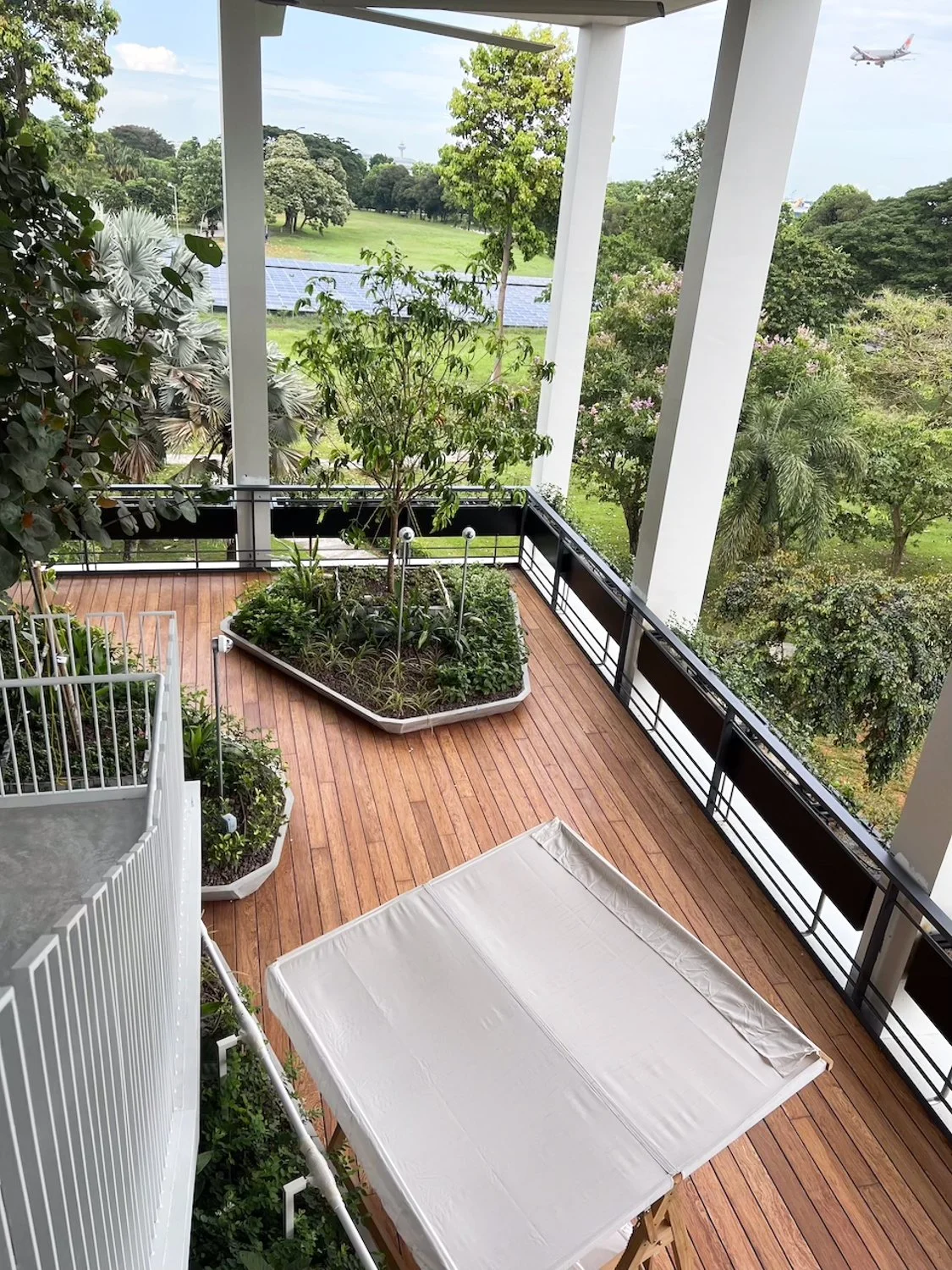 View of a balcony with a wooden floor, white railing, and a small garden bed with a tree and plants. Beyond the balcony, there are trees, open green space, solar panels, and an airplane flying in the sky.