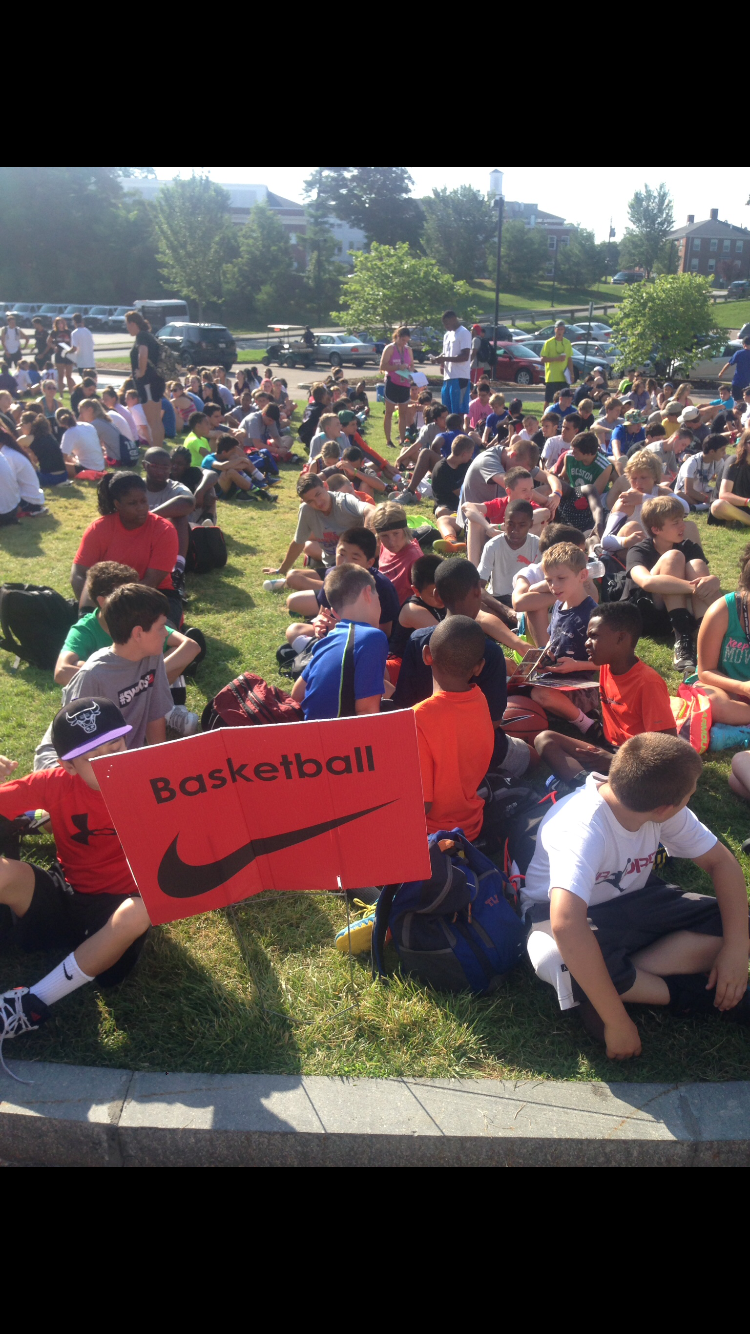 A large group of children sitting on the grass outside, with some adults standing nearby, during daytime. A red sign with the words 'Basketball' and a Nike logo is in the foreground.