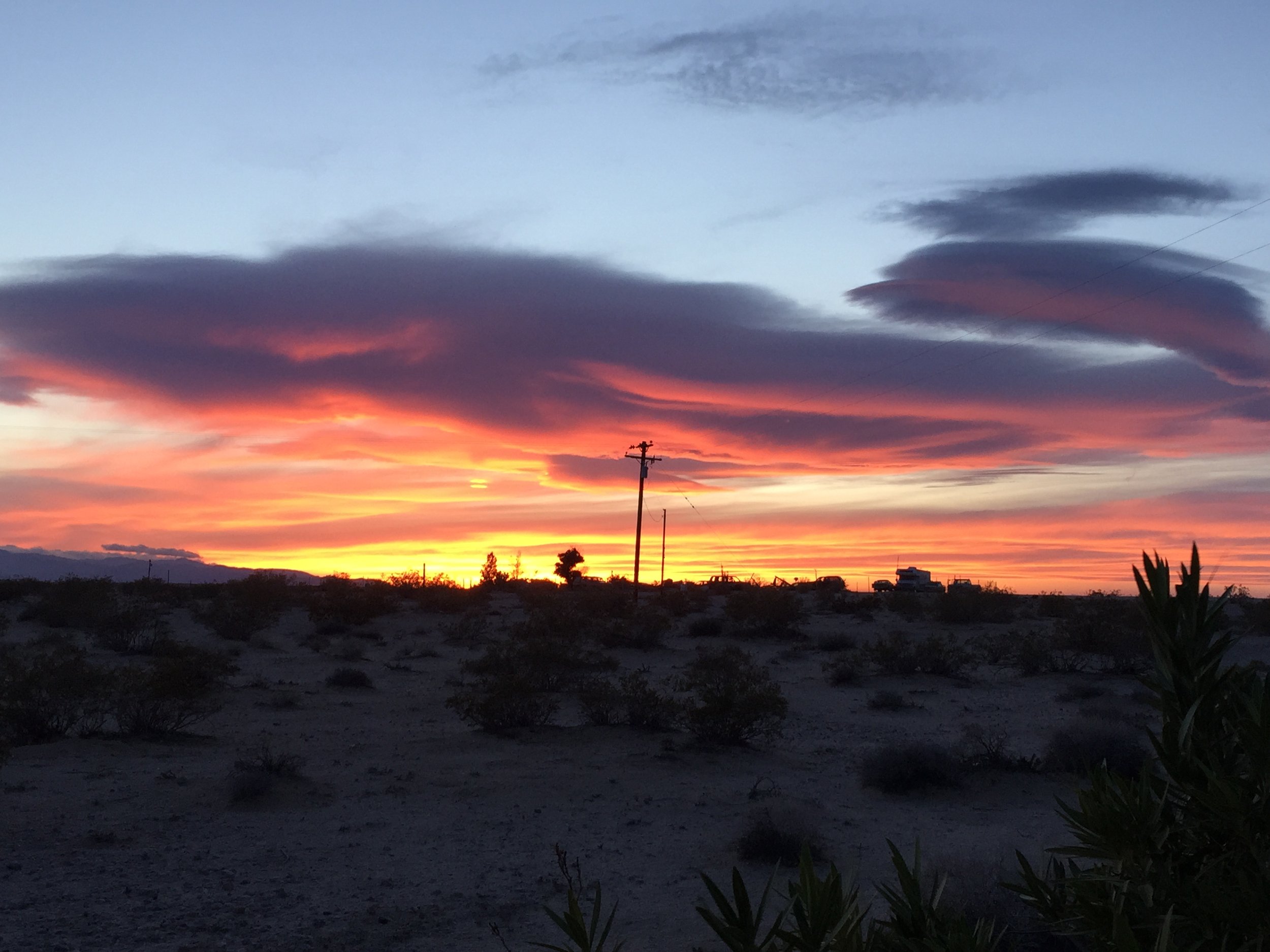 Sunset over a desert landscape with sparse vegetation, electrical poles, and a few buildings on the horizon, under a sky with dark purple and pink clouds.