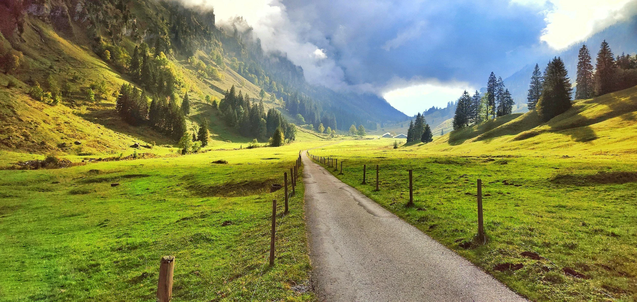A narrow road leading through a lush green valley with rolling hills and scattered pine trees, surrounded by mountains under a partly cloudy sky.