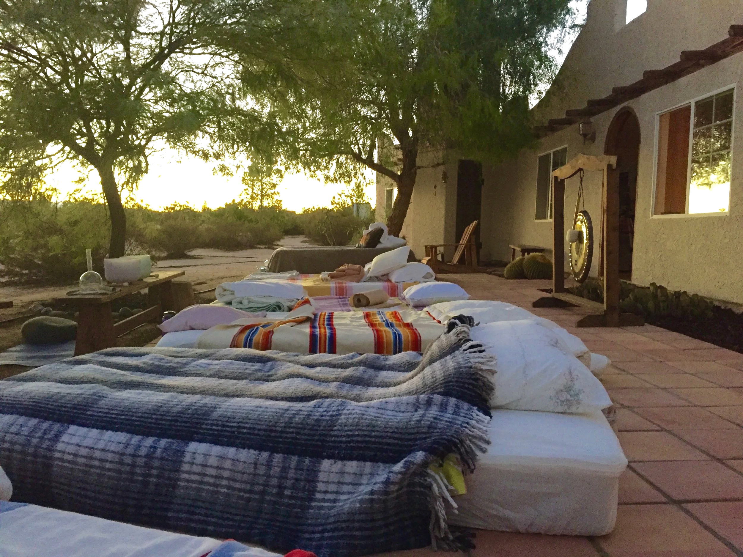 Outdoor patio scene at sunset with mattresses, blankets, and pillows arranged on the ground, and a musical gong on a stand near the wall of a house.