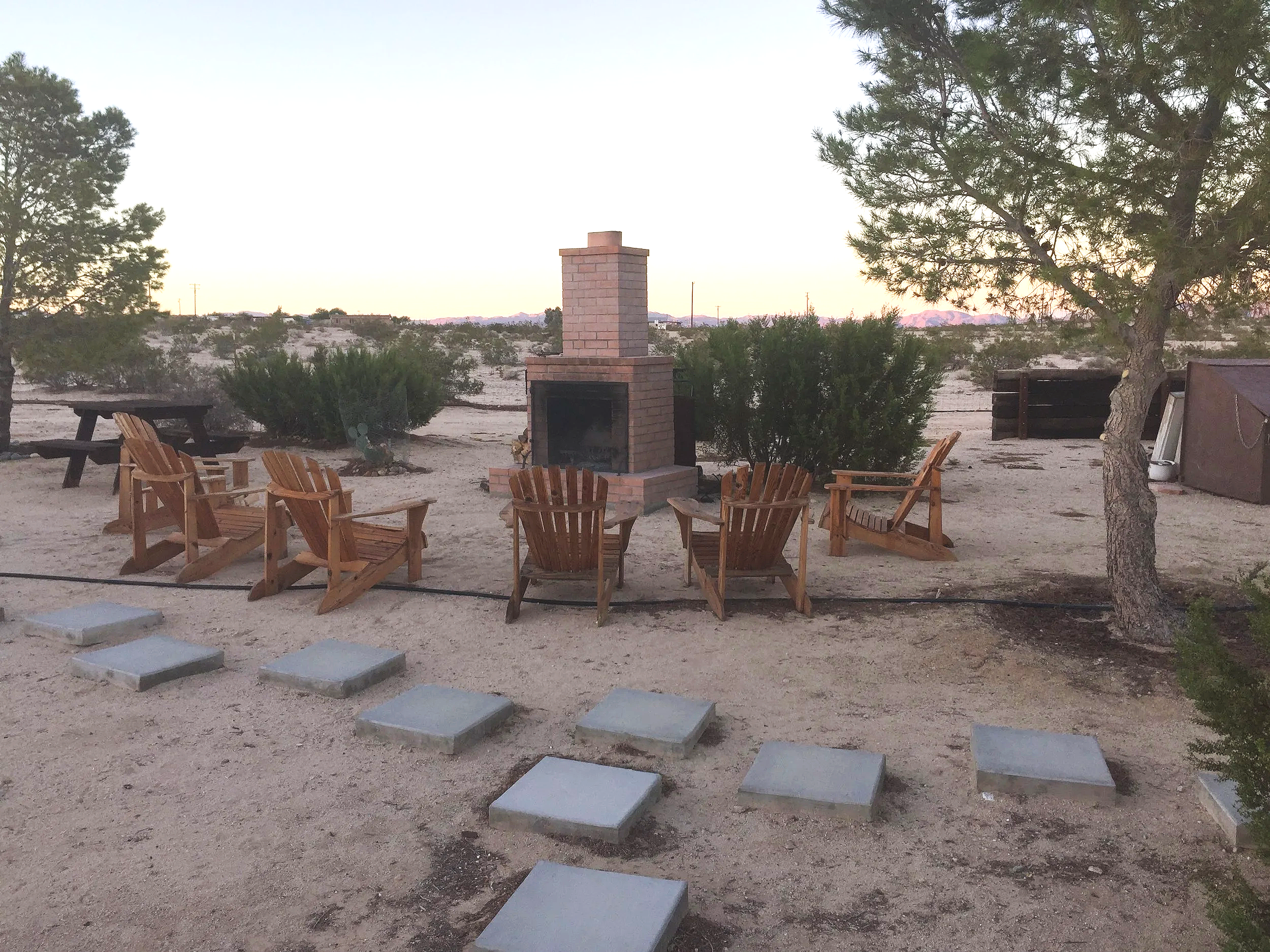 Outdoor desert patio with six wooden Adirondack chairs arranged in a semi-circle around a brick outdoor fireplace, surrounded by desert plants and trees, with a mountain range in the background during sunset.