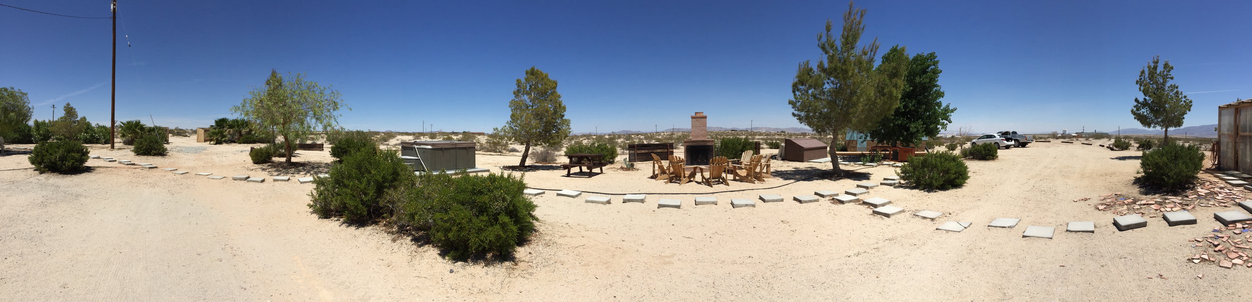Panoramic view of a desert outdoor space with gravel ground, scattered bushes and trees, a firepit with chairs around it, a few parked cars, and a clear blue sky.