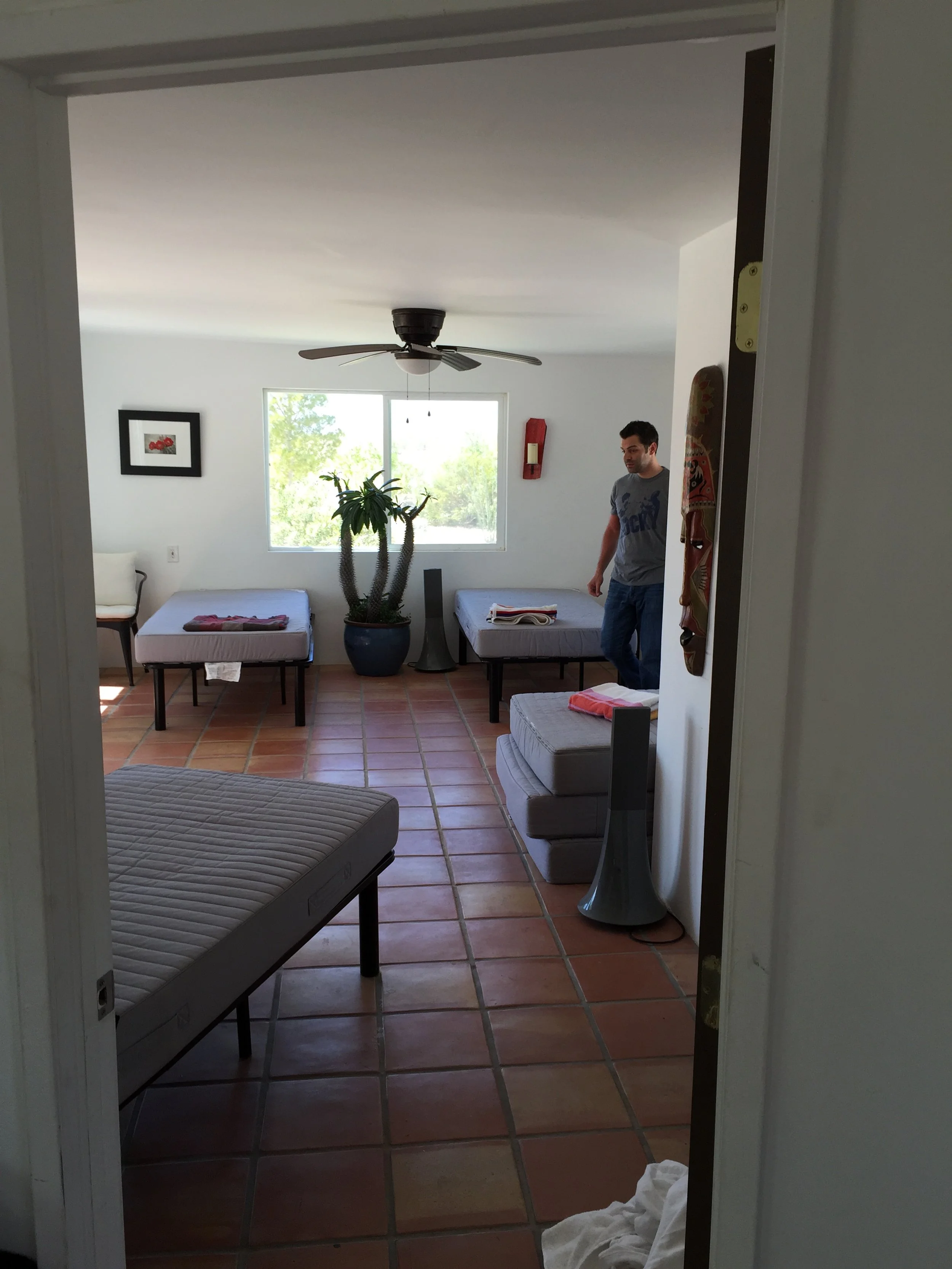 A bedroom with terracotta tile flooring, two beds, large window, ceiling fan, potted plant, and a man standing near one of the beds.