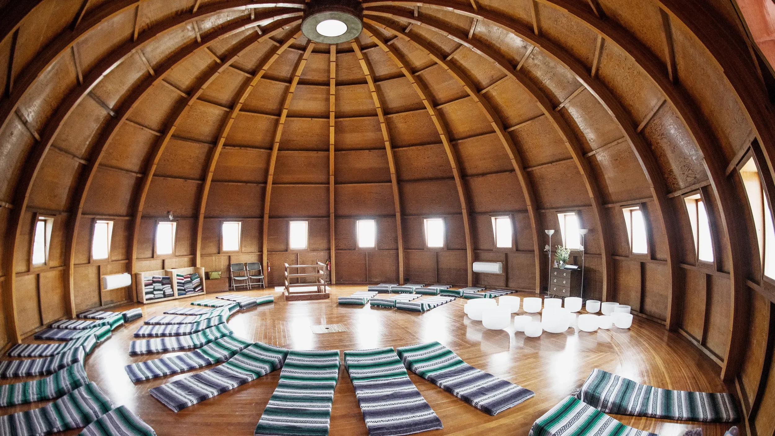 Inside a round wooden domed room with multiple small windows, arranged with yoga mats and cushions on a wooden floor, and some white glowing chairs.