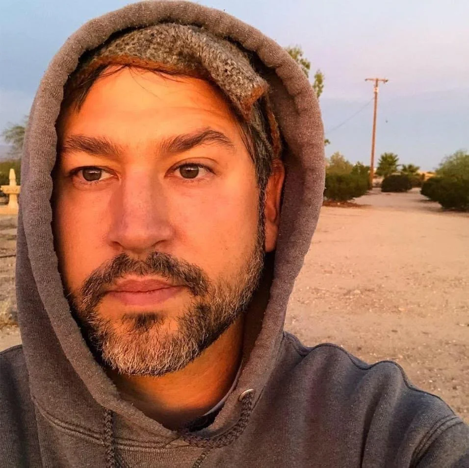 Close-up of a man with a beard wearing a gray hoodie, outdoors in a desert-like area during sunset.