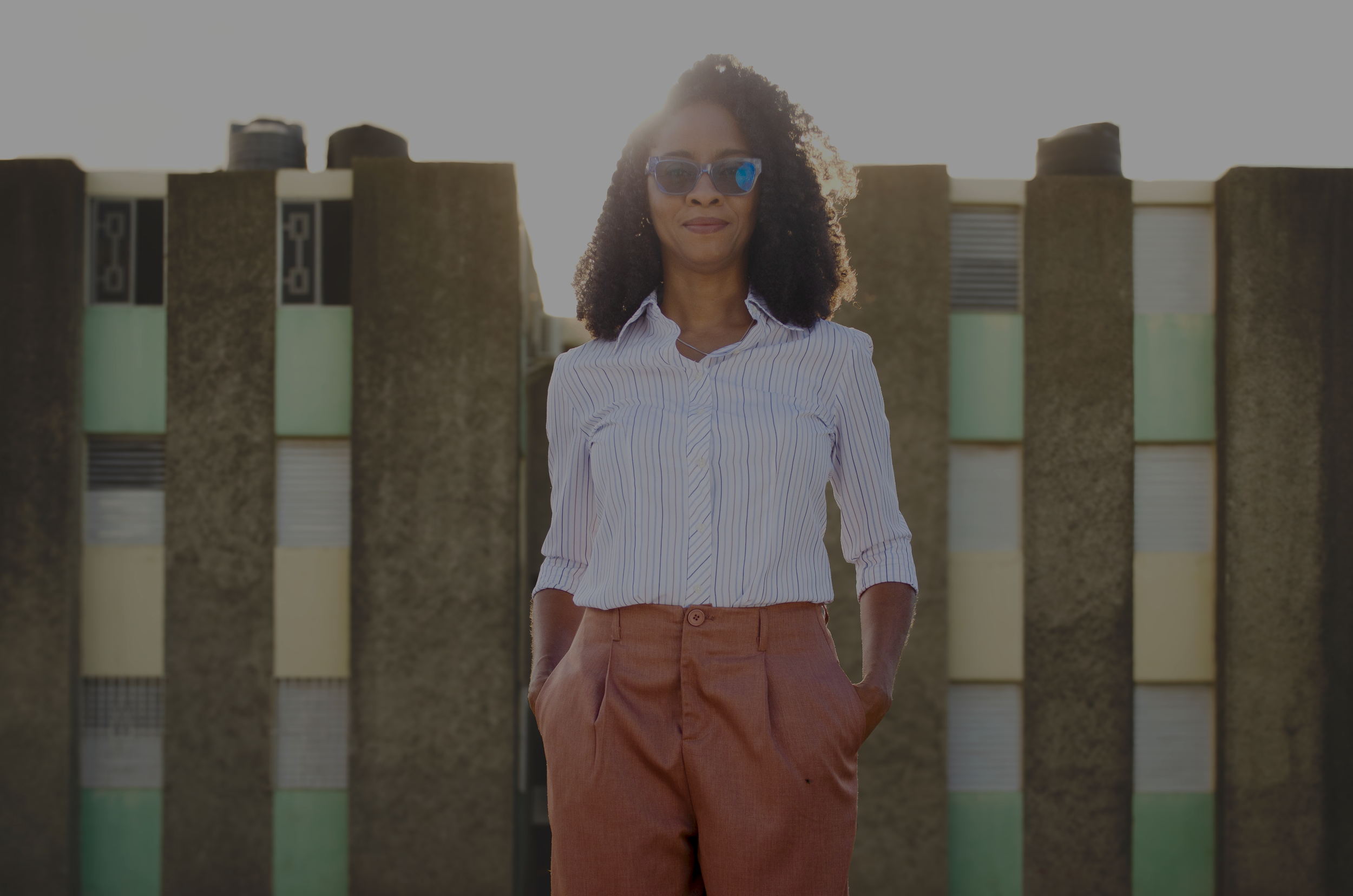 A woman with curly hair wearing sunglasses, a white striped shirt, and rust-colored pants standing outdoors with a building and sunlight in the background.