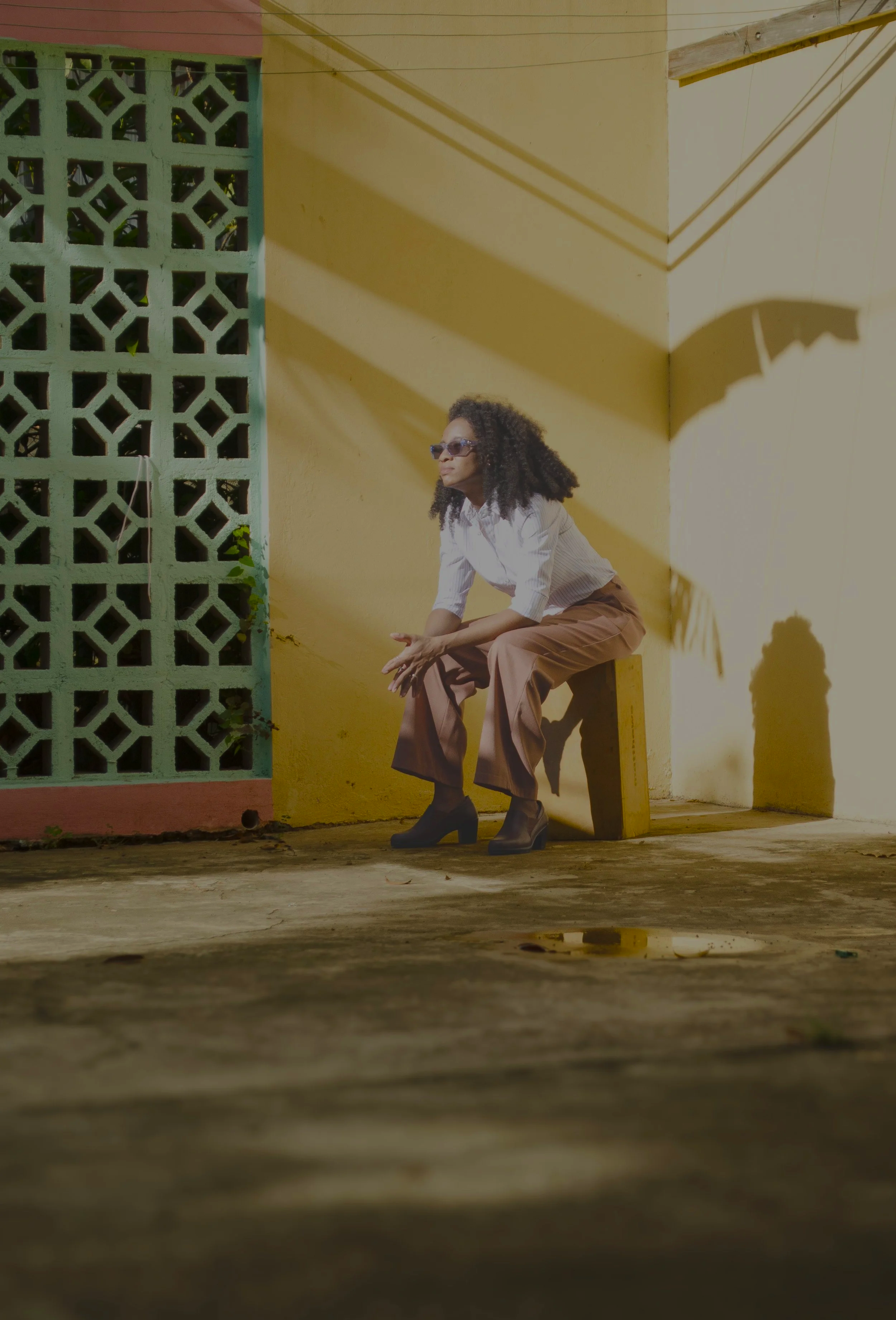 A woman with curly hair wearing sunglasses, a white blouse, wide-legged brown pants, and black high heels, sitting on a small wooden stool against a yellow wall with a green decorative brick pattern on an outdoor patio.