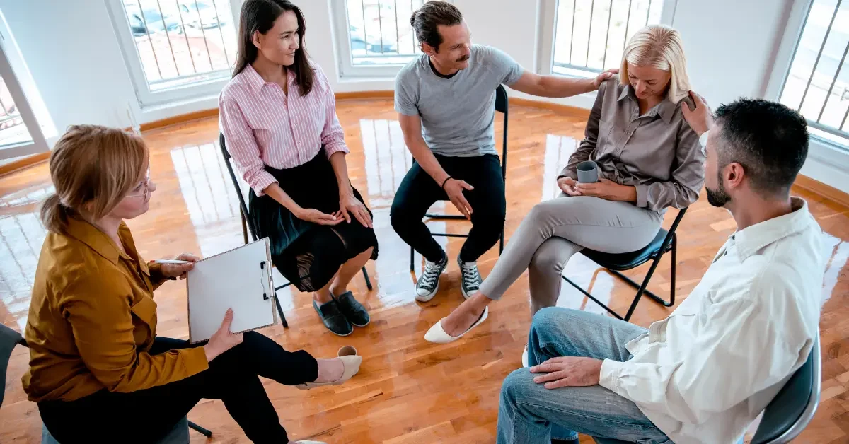 Group of people participating in a therapy or support group session in a bright room with large windows, sitting in a circle and engaging in conversation.