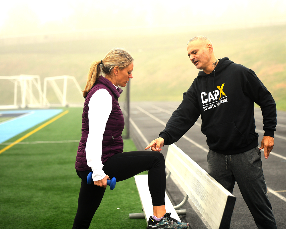 A woman is performing a step-up exercise with a dumbbell while a trainer points at her knee in an outdoor track and field setting with trainer Christian Caputo