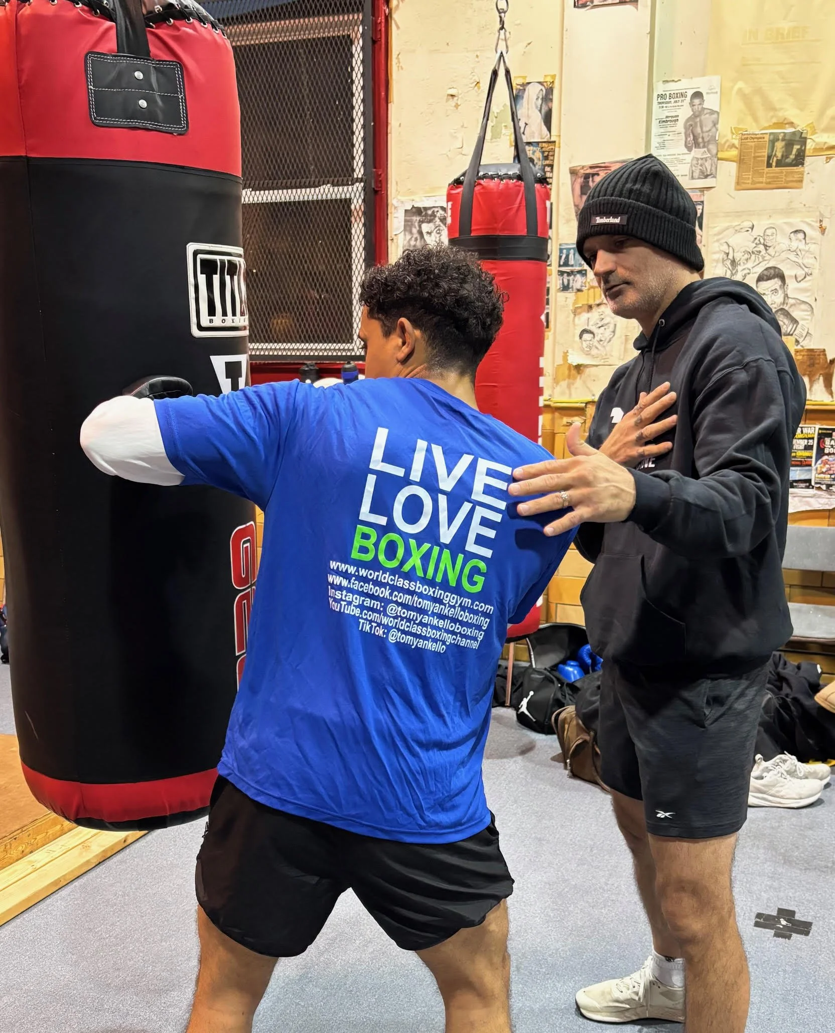 A boxing trainer coaching a boxer in a gym, demonstrating a punching technique near a heavy punching bag, with posters on the wall and gym equipment visible.
