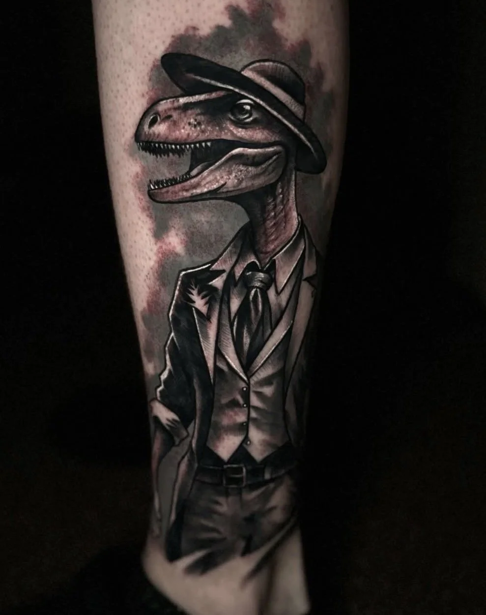 Black and grey tattoo of a dinosaur wearing a suit and hat on the arm, completed by Cody Eckenrode at CREate Tattoo Studio.