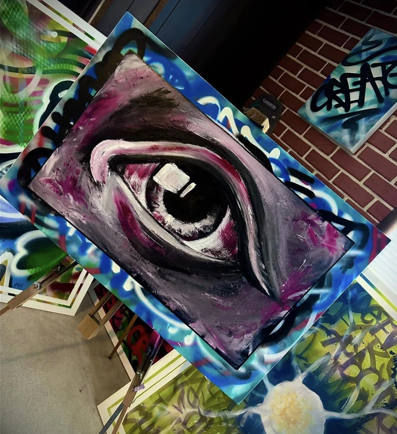 A painting of a large eye with black, white, pink, and purple hues, displayed on an easel in an art gallery with other graffiti-style artwork around made by Cody Eckenrode