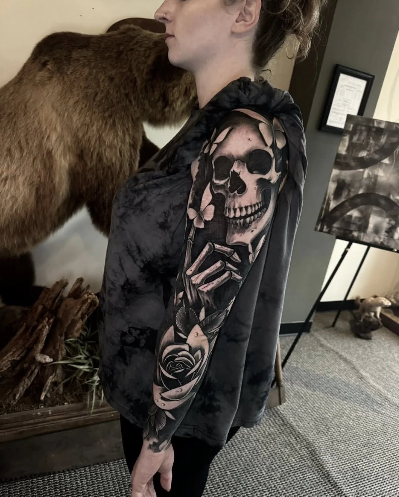 A person with a large tattoo sleeve featuring a skull and rose designs, standing next to a bear prop or statue, in an indoor space with artwork and decor completed by Cody Eckenrode at CREate Tattoo Studio.
