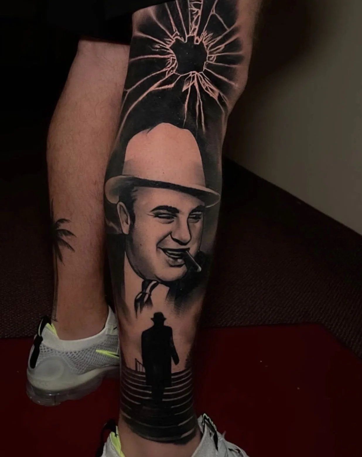High-contrast black and grey tattoo of Al Capone smoking a cigar, framed under a shadowed silhouette with a broken window from a bullet impact by Codo Eckenrode at CREate Tattoo Studio.