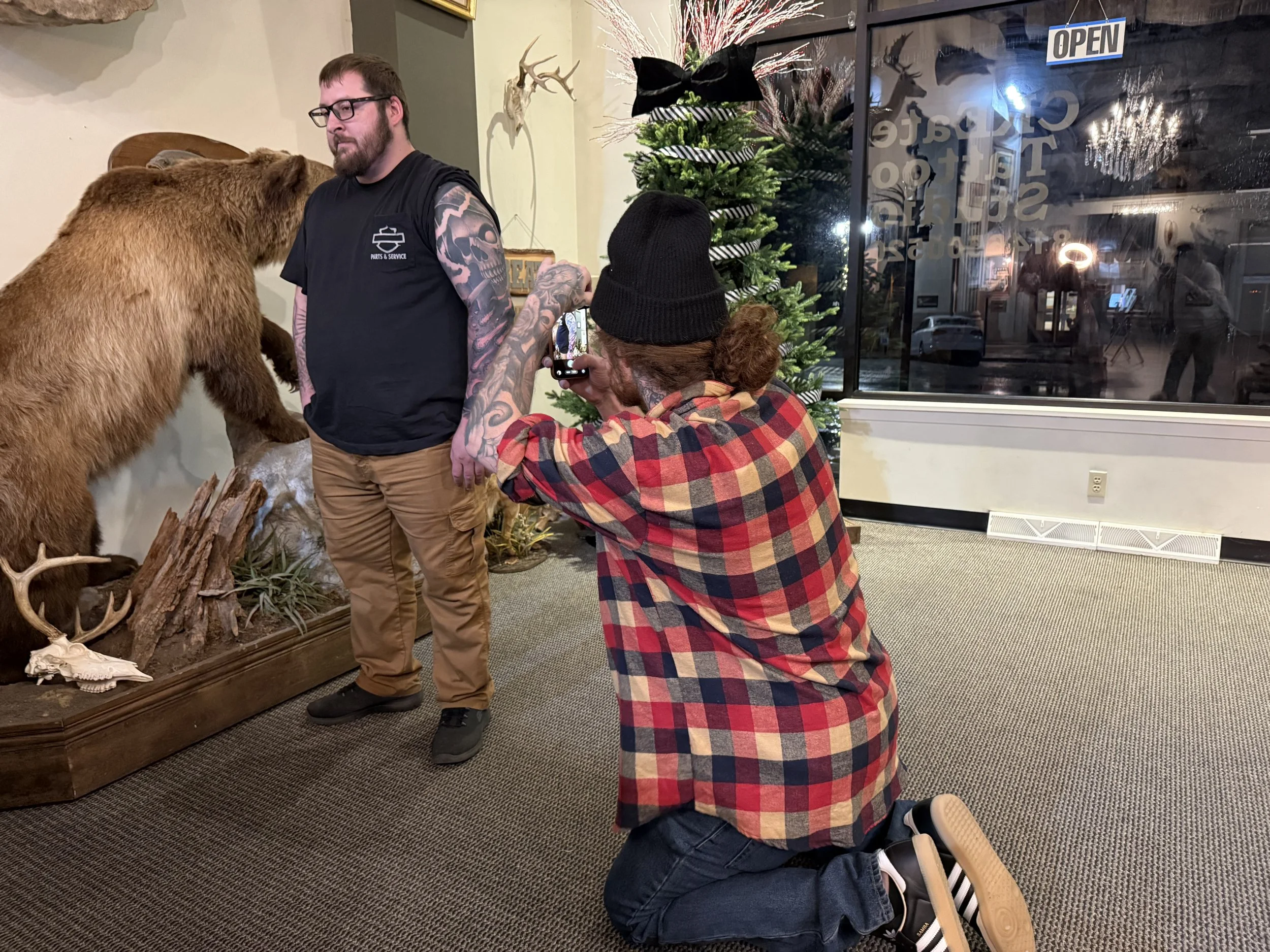 Cody Eckenrode kneeling to photograph a freshly completed tattoo on a client’s lower arm at CREate Tattoo Studio.