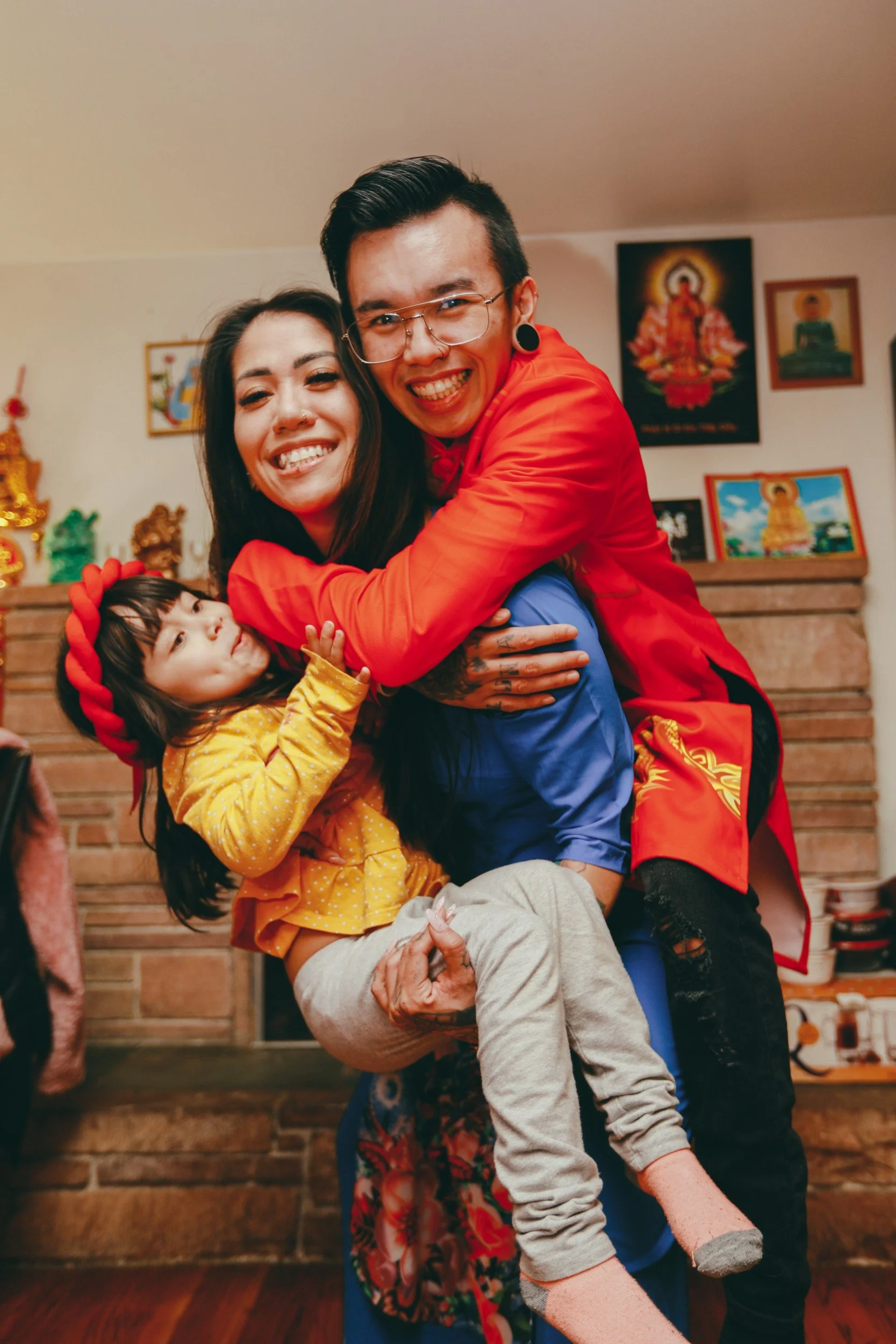 Three people embrace and smile, a man, woman, and young girl, in a warm home decorated with religious and holiday artwork.