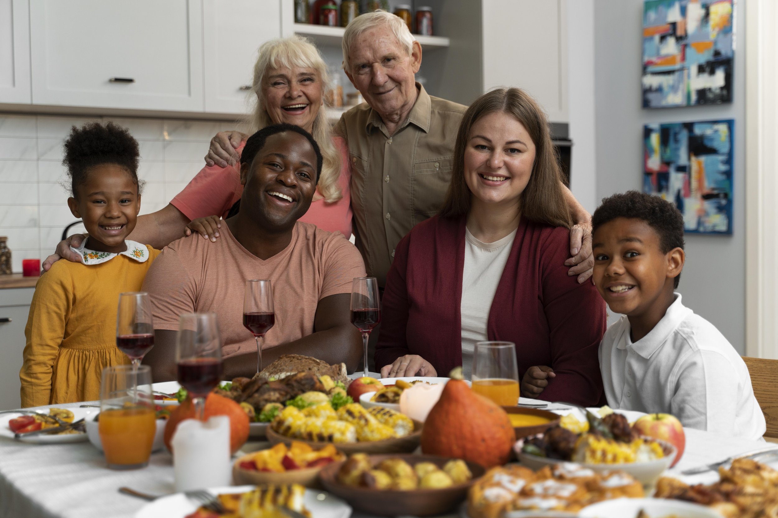 A happy multi-generational family gathered around a Thanksgiving feast table, smiling and posing for the camera in a modern kitchen.