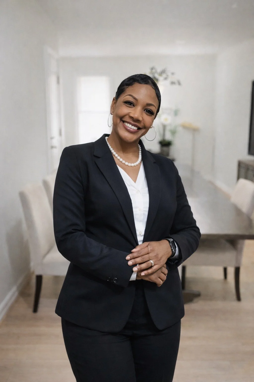 A smiling woman in a black blazer and pearl necklace standing in a well-lit room with a dining table and chairs in the background.