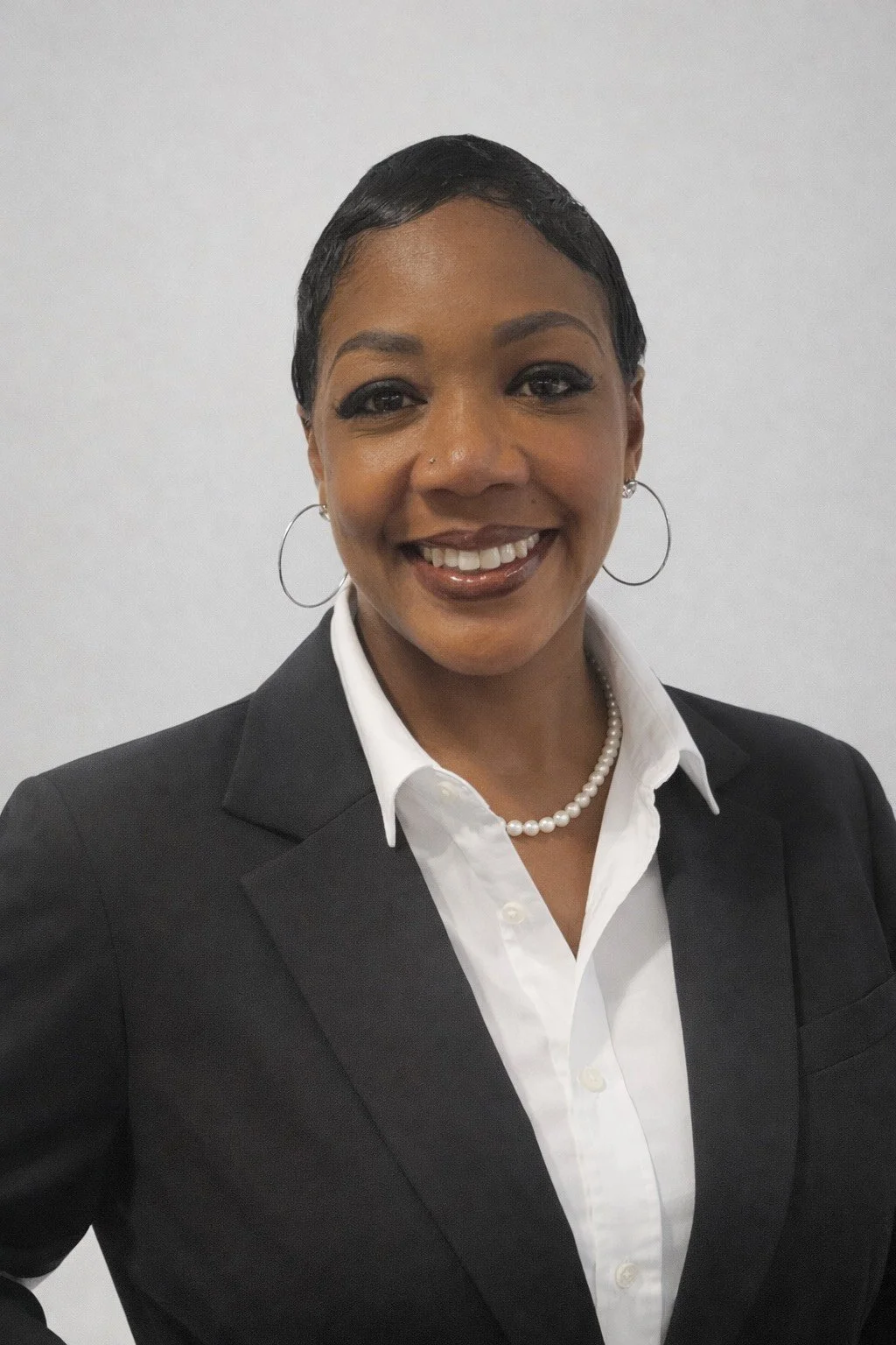 A smiling woman in a black blazer and white shirt, wearing pearl jewelry and hoop earrings, against a plain light background.
