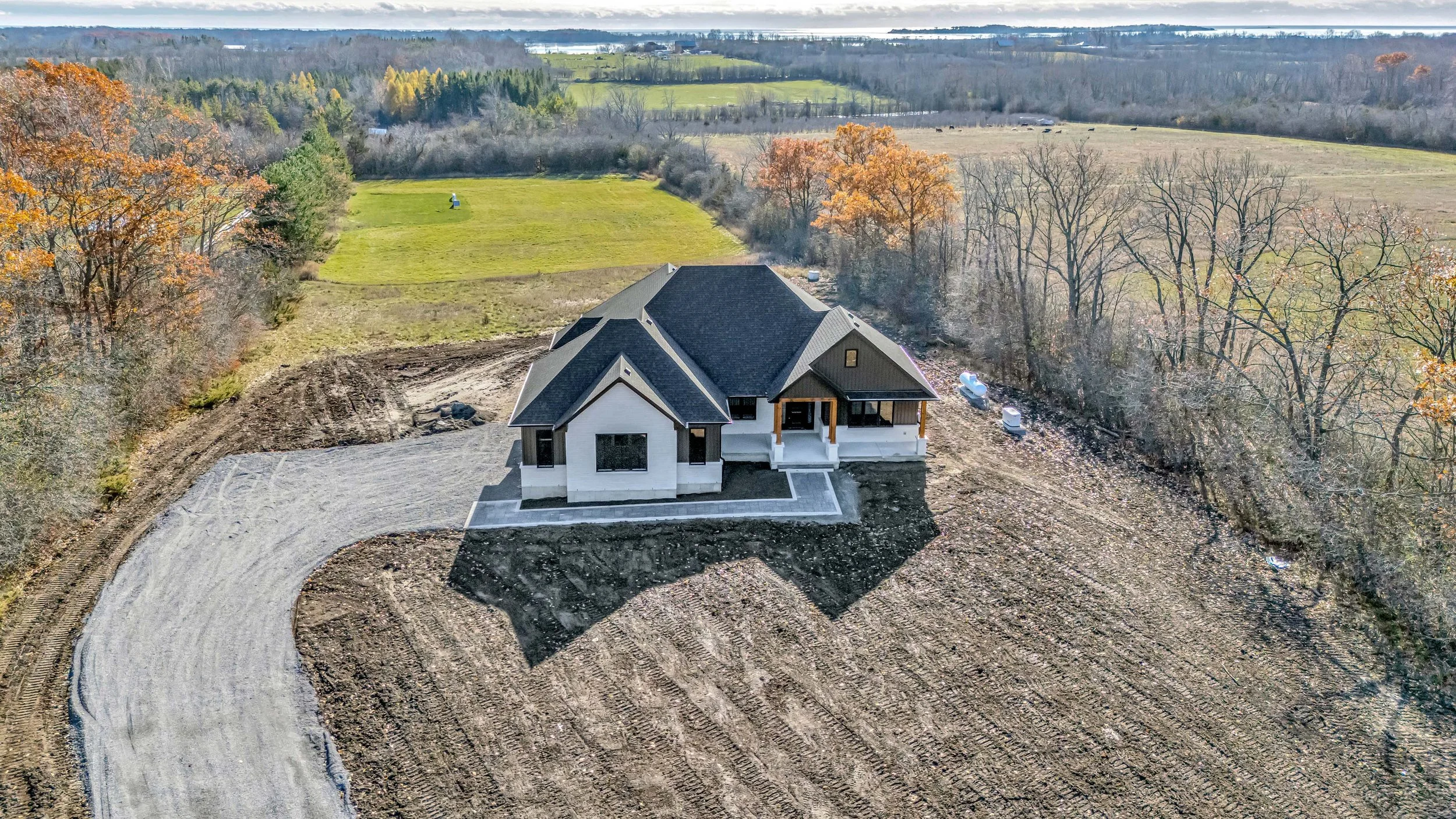 A house under construction in a rural area, surrounded by trees, with a curved gravel driveway and a large grassy field in the background.