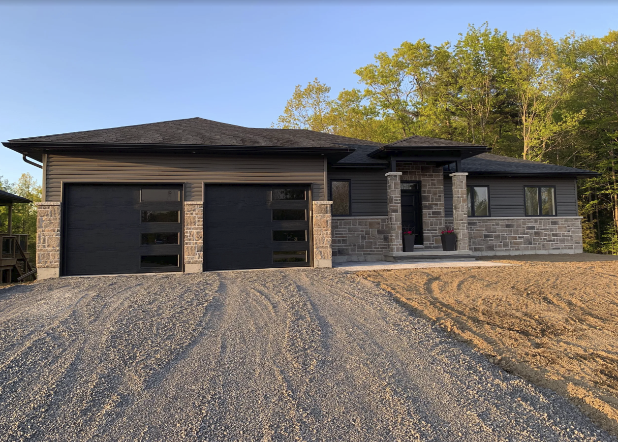 Modern house with stone facade, black garage doors, and front porch with two columns and potted plants, surrounded by trees.