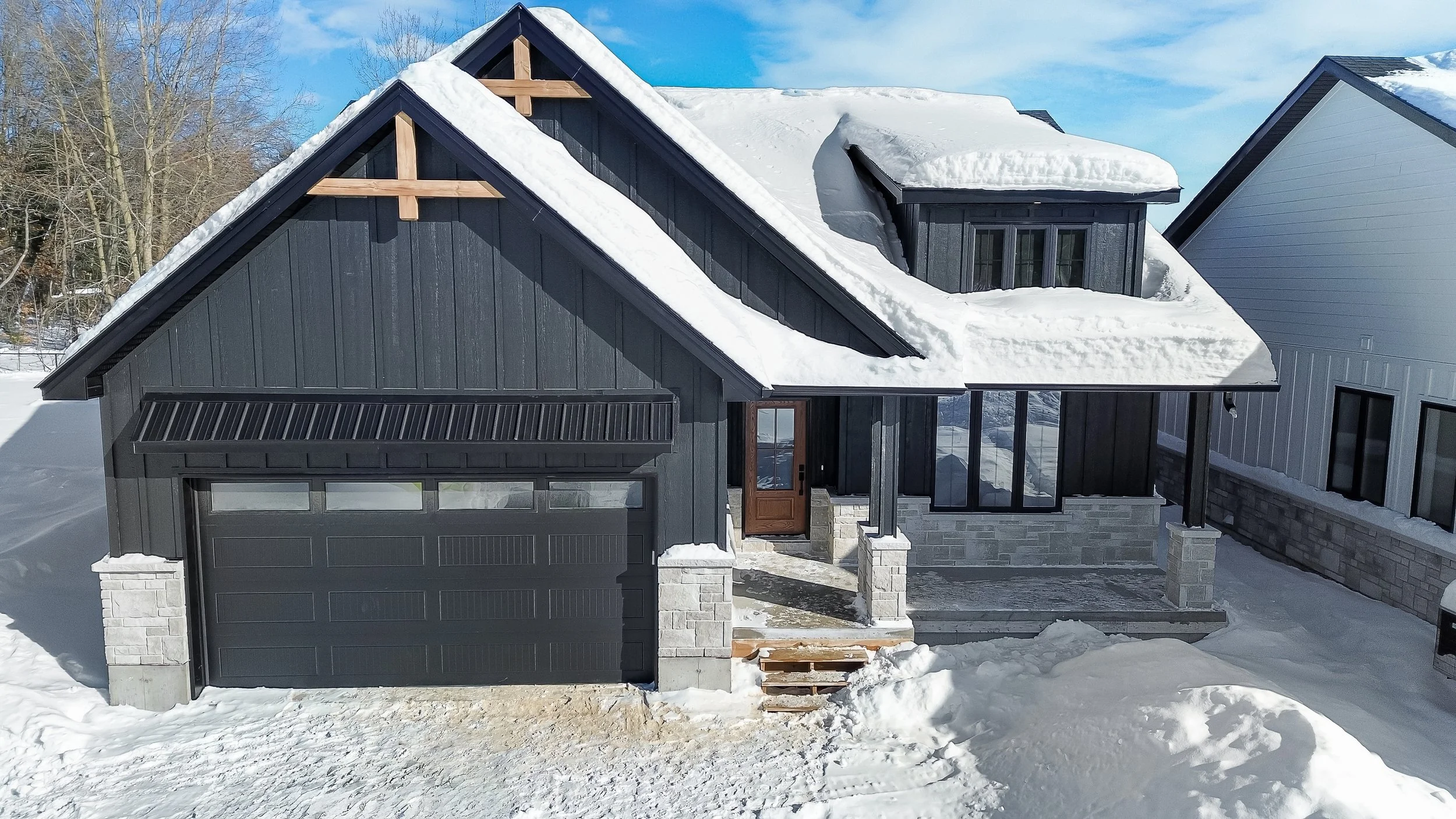 A modern house with a black exterior, stone accents, and a sloped roof covered with snow. The house has a front porch and a garage door, with a snowy driveway leading up to it.