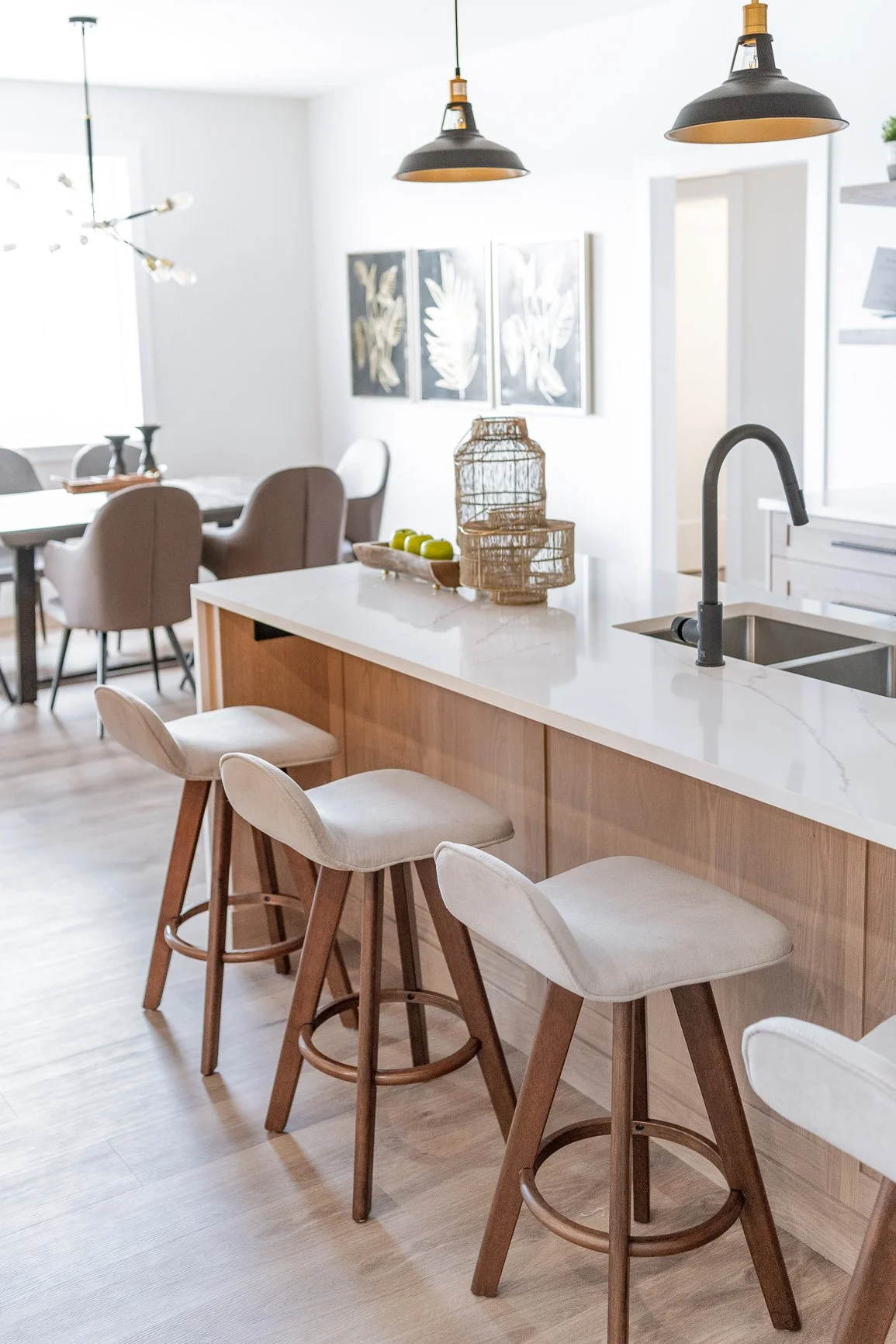 Modern kitchen with four barstools at a white marble island, black pendant lights, and artwork on the wall, with a dining area visible in the background.
