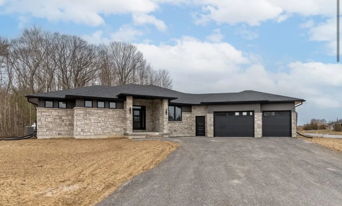 Modern house with stone and black siding exterior, two garage doors, a gravel driveway, and leafless trees in the background under a partly cloudy sky.