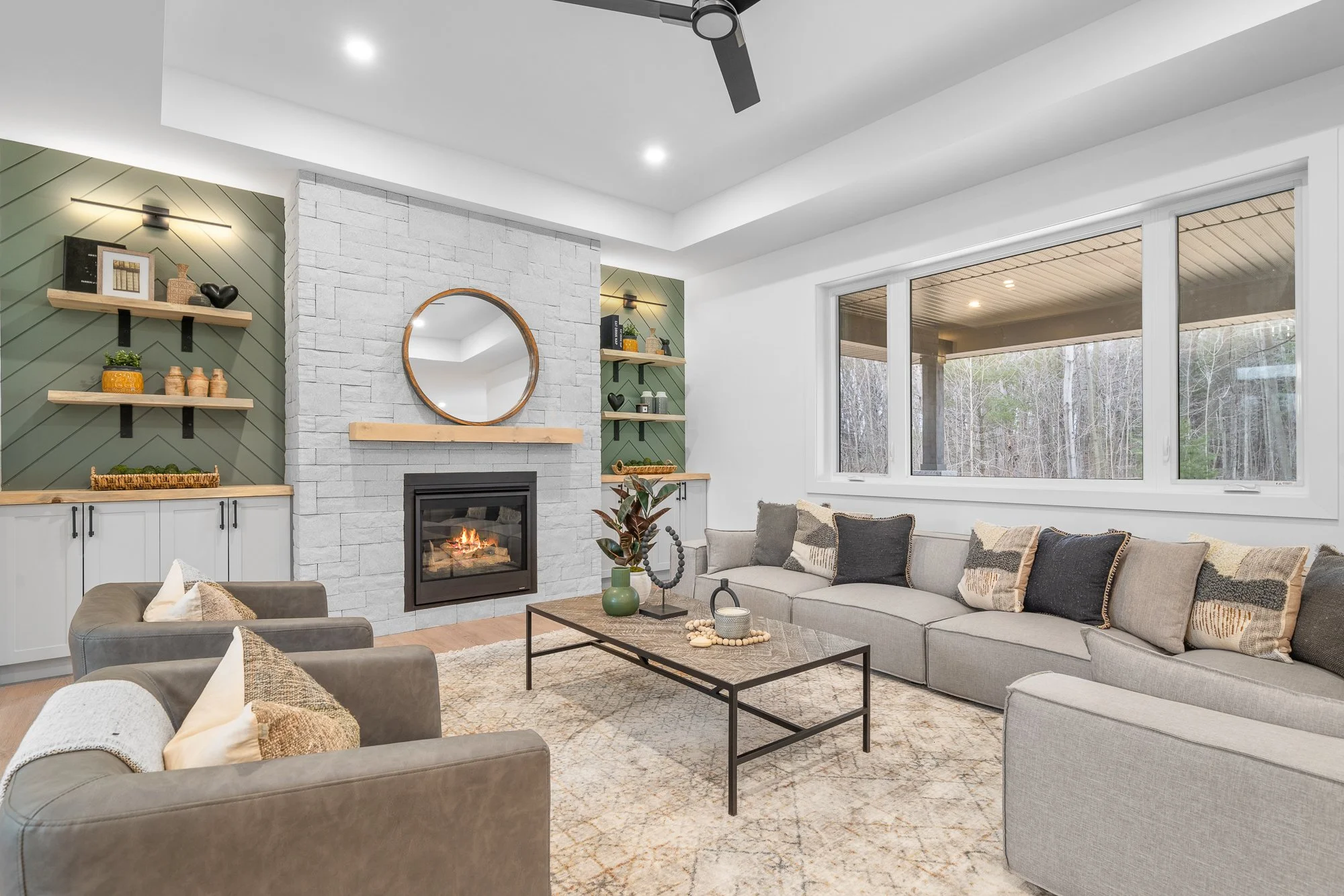 Living room with a large sectional sofa, two armchairs, a coffee table, a fireplace with a mirror above, and large windows showing a wooded area outside.