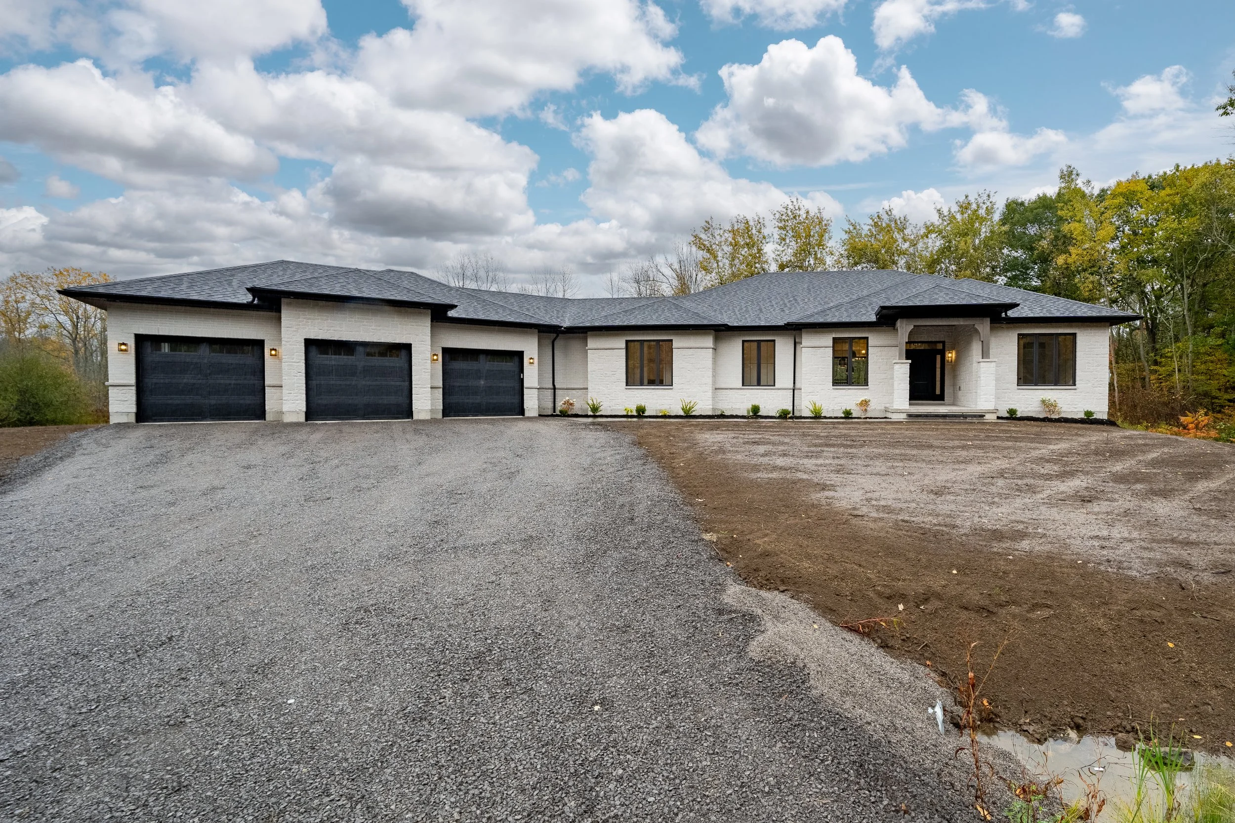 Modern white brick house with black garage doors, black roof, and front porch, surrounded by a gravel driveway and newly planted bushes, with trees and a cloudy sky in the background.