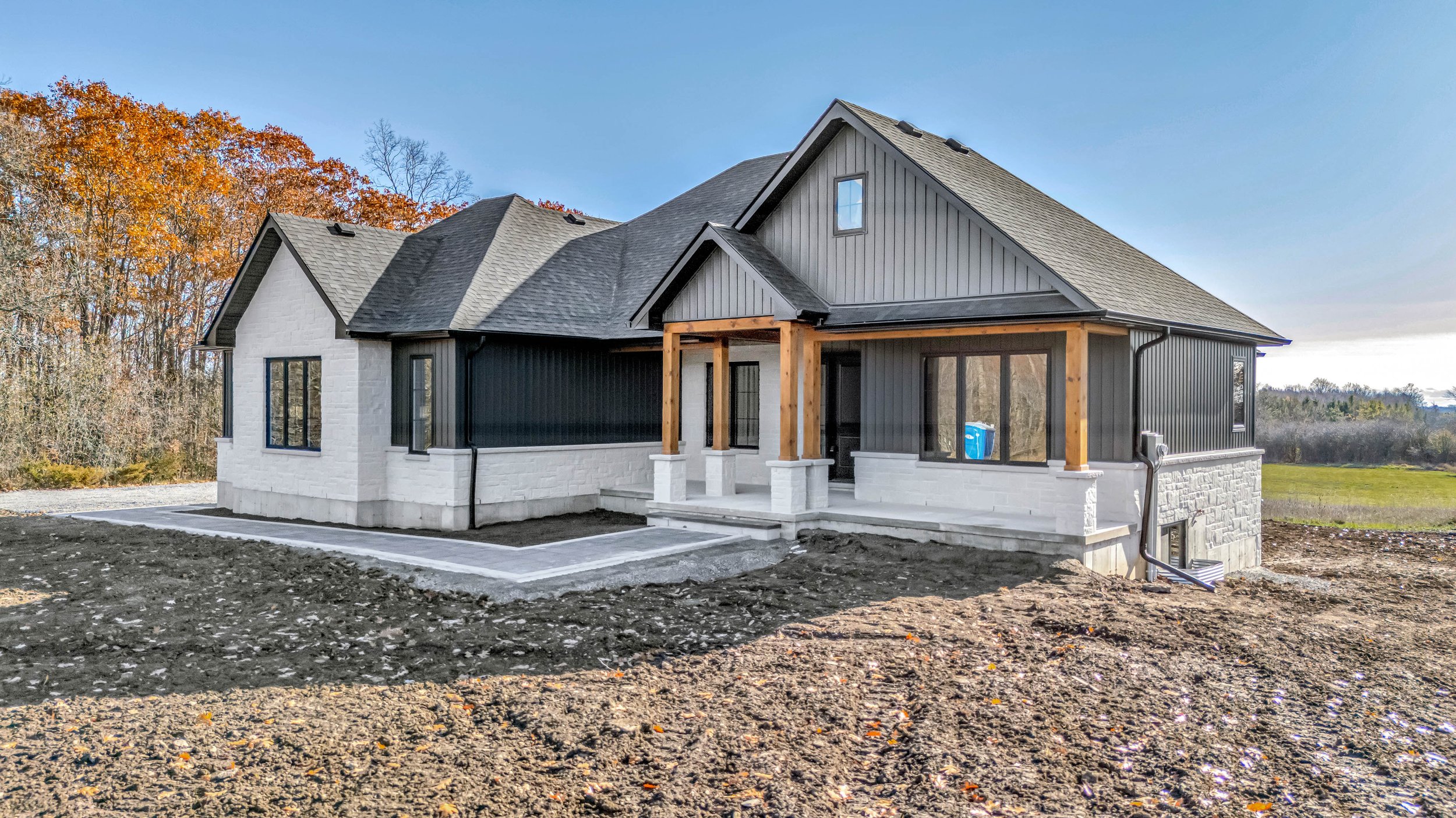 A house under construction with a gray roof, black and white exterior walls, and wooden porch columns, surrounded by bare ground and a backdrop of trees with fall foliage.