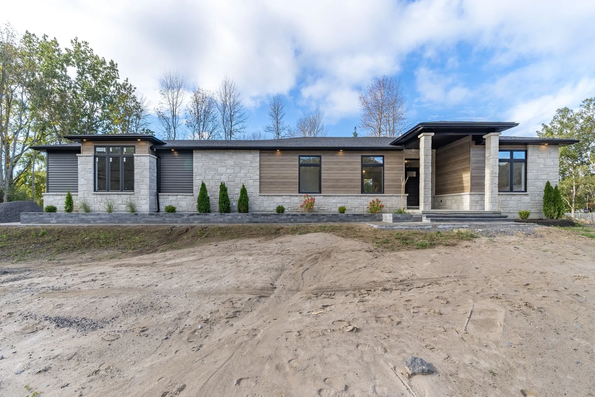 Modern house with stone and wooden exterior, black windows, and a covered entrance. The front yard is still under construction with dirt and rocks, and a row of small shrubs lines the house.