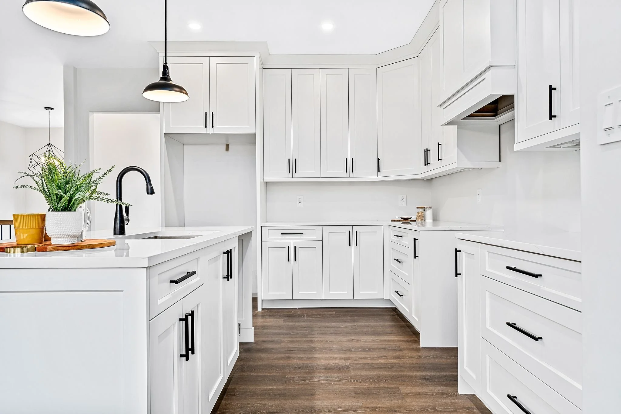Modern white kitchen with black handles, dark wood floor, plants, and pendant lights.