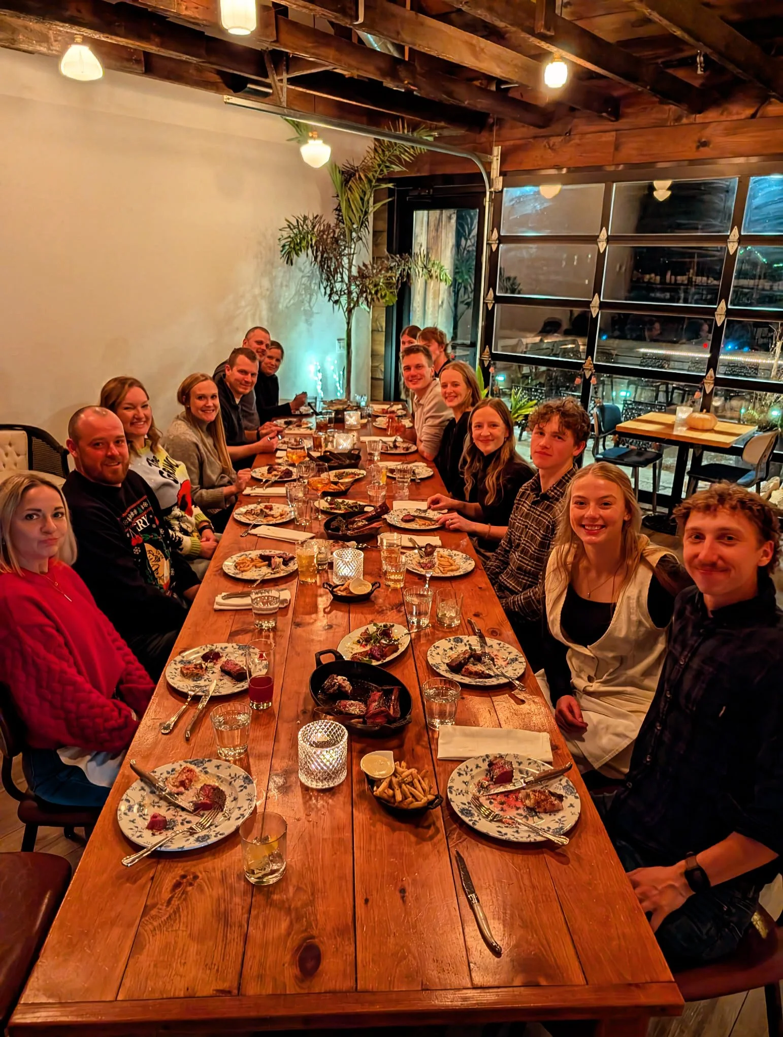 A large group of people sitting around a long wooden dining table enjoying a meal in a cozy restaurant with warm lighting and large windows.