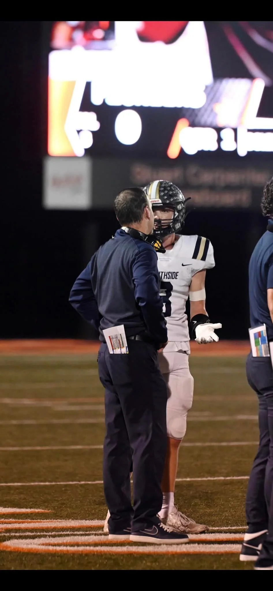 A football player in a white uniform with number 3 talking to a coach in a dark jacket on the field at night, with other staff and a scoreboard in the background.