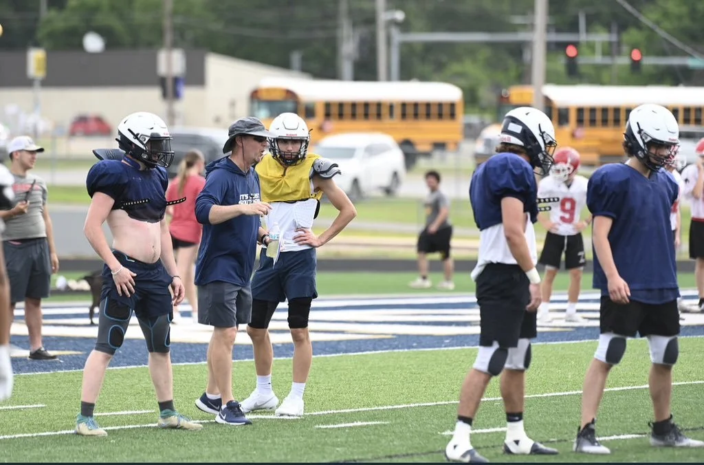 Youth football players wearing helmets and gear on a field, with a coach or trainer giving instructions, spectators and school buses in the background.