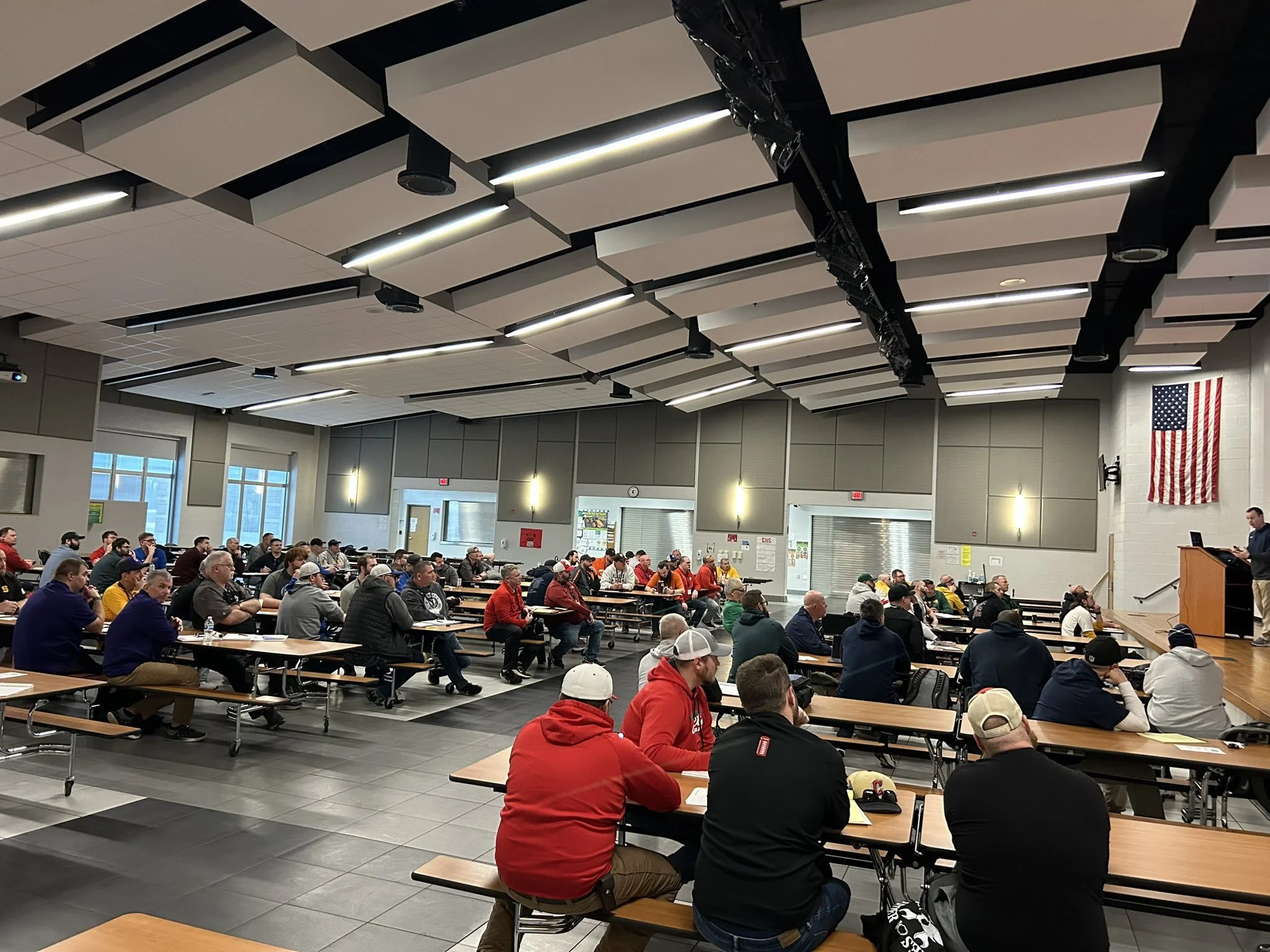 A large group of people seated at tables in a spacious, well-lit cafeteria or conference room, listening to a speaker on stage with a podium, an American flag hanging on the wall.