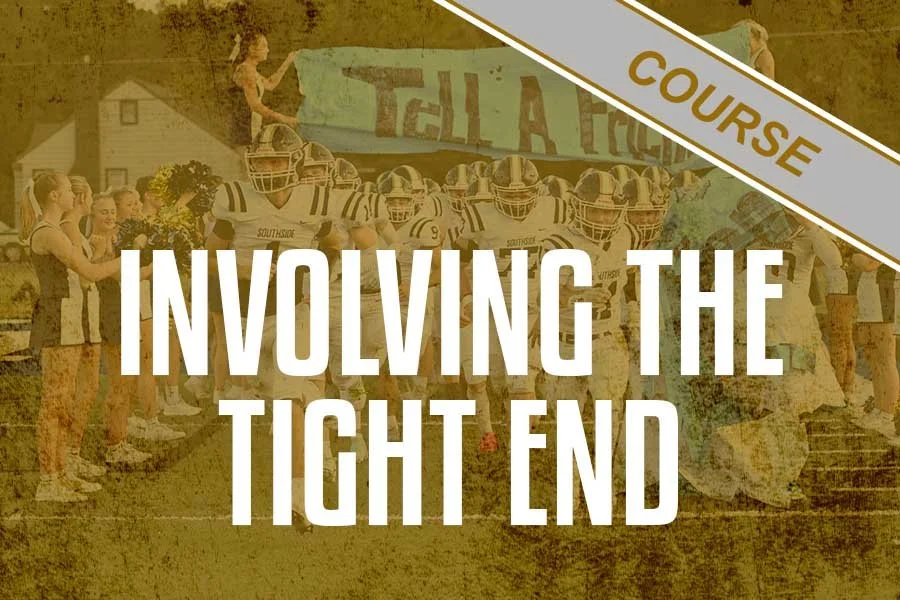 A group of football players lined up on the field, with cheerleaders and spectators surrounding them, and a banner in the background.